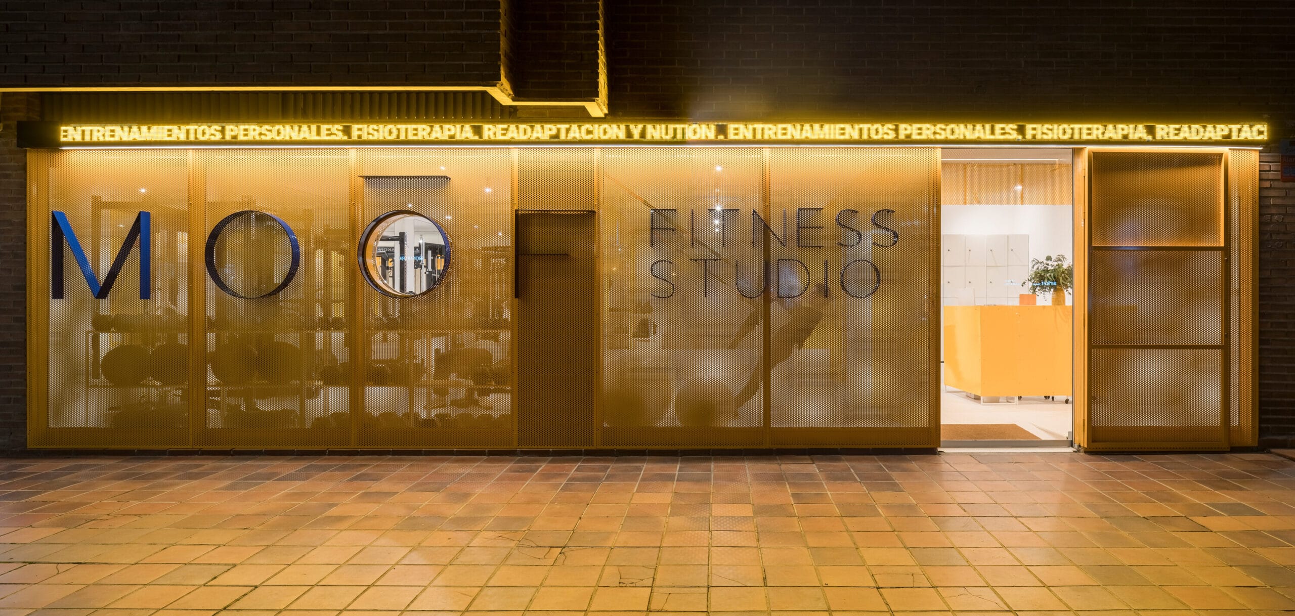Gevel van MOO Fitness Studio met een goudkleurige geperforeerde wand en verlichte tekst over training en fysiotherapie