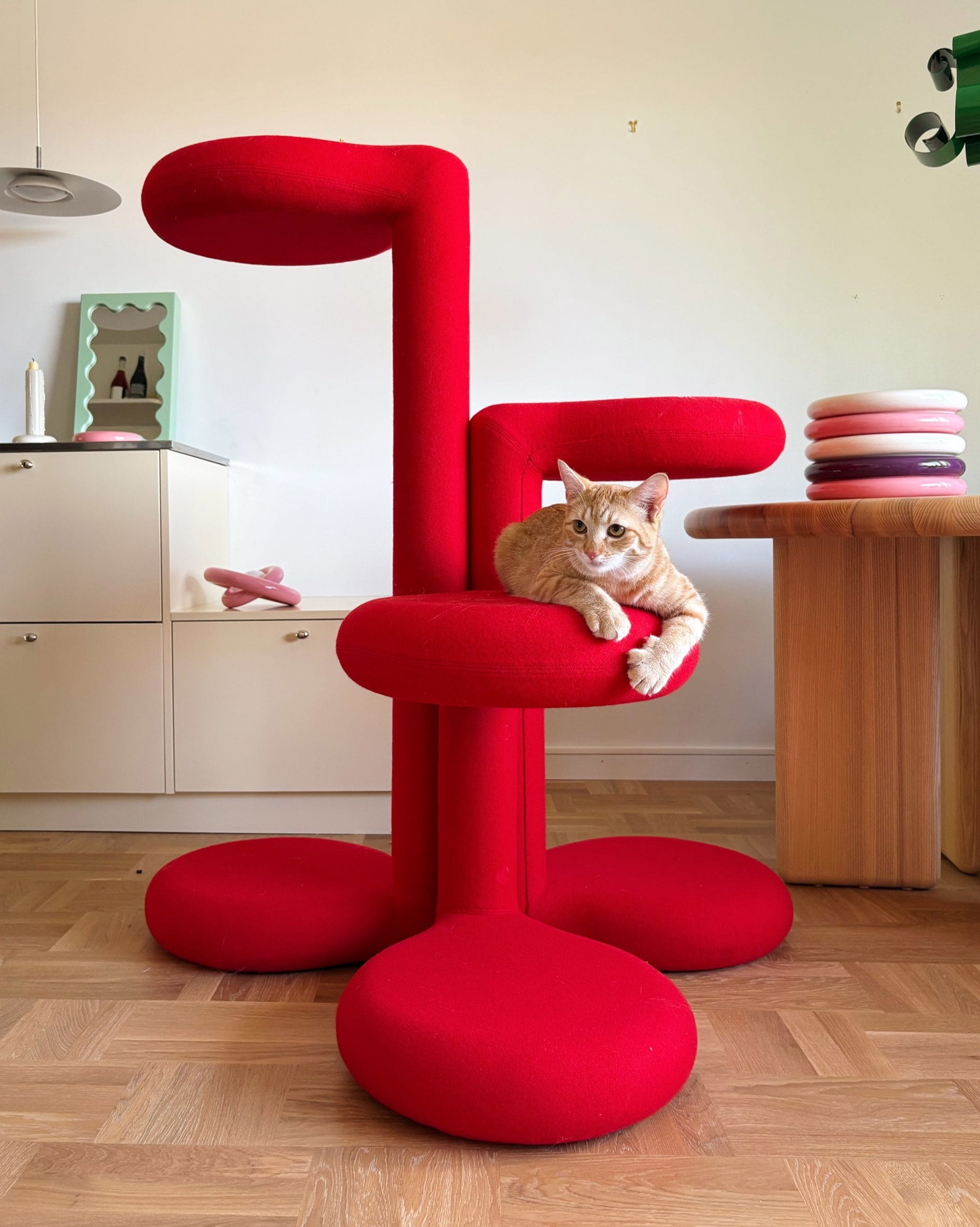 Een oranje kat ligt op een moderne, felrode krabpaal met ronde plateaus in een licht interieur