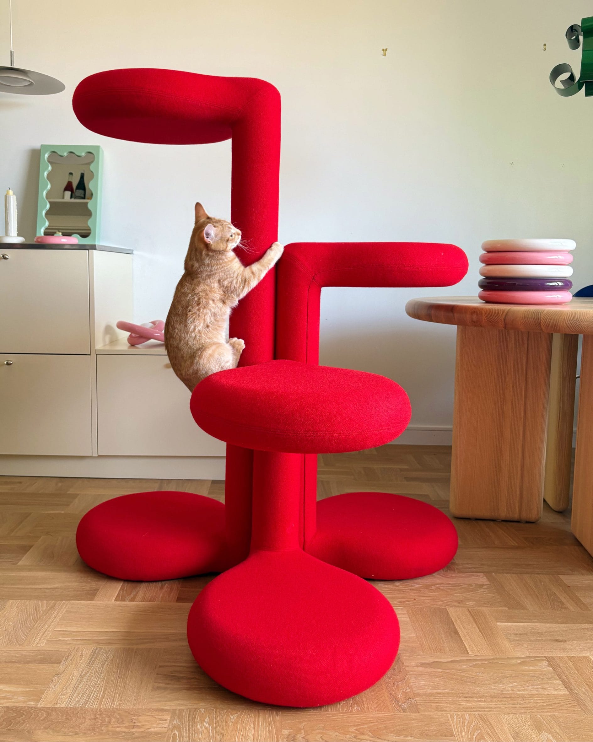 Een oranje kat klimt op een moderne, felrode krabpaal met ronde plateaus in een minimalistisch interieur