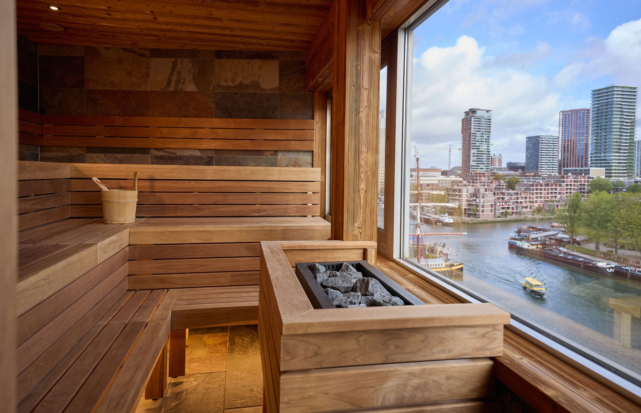 Houten sauna met een groot raam dat uitkijkt over een rivier en de skyline van een stad met moderne hoogbouw