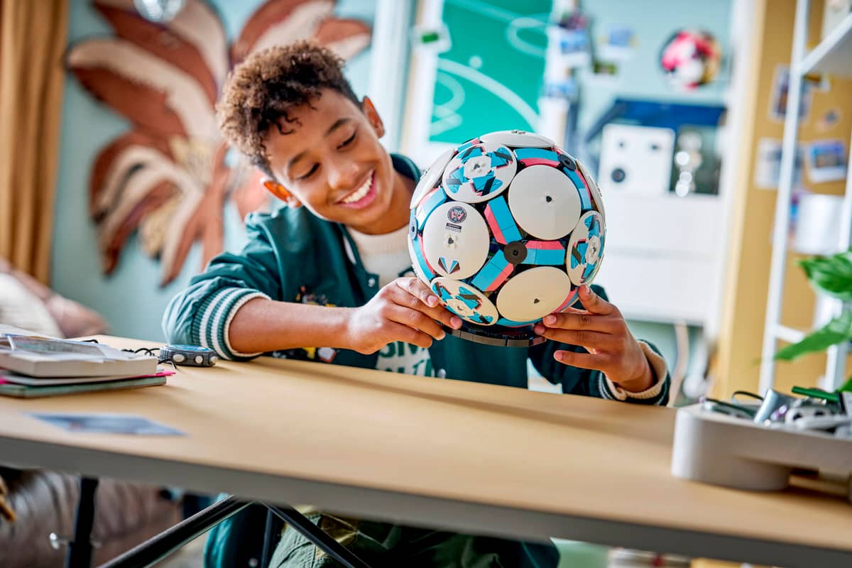 Een lachende jongen aan een bureau bewondert een zelfgebouwde, kleurrijke LEGO-voetbal