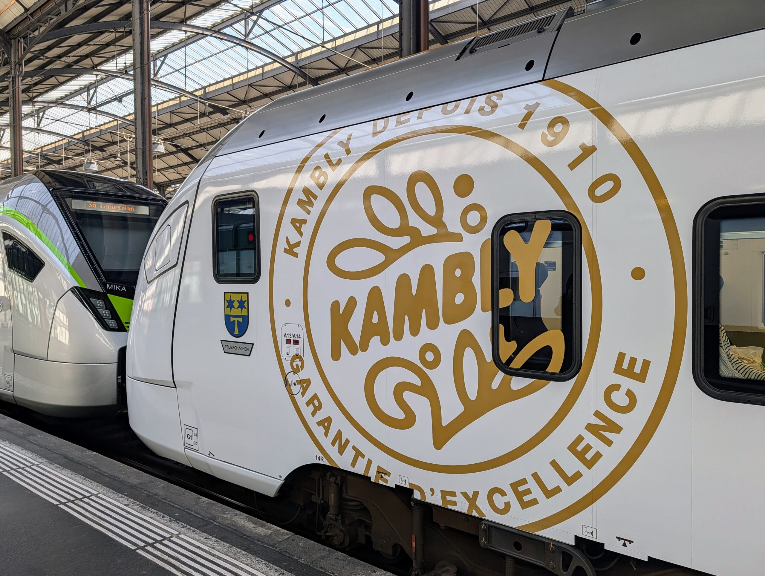Zijaanzicht van een witte trein met een groot goudkleurig Kambly-logo op een station