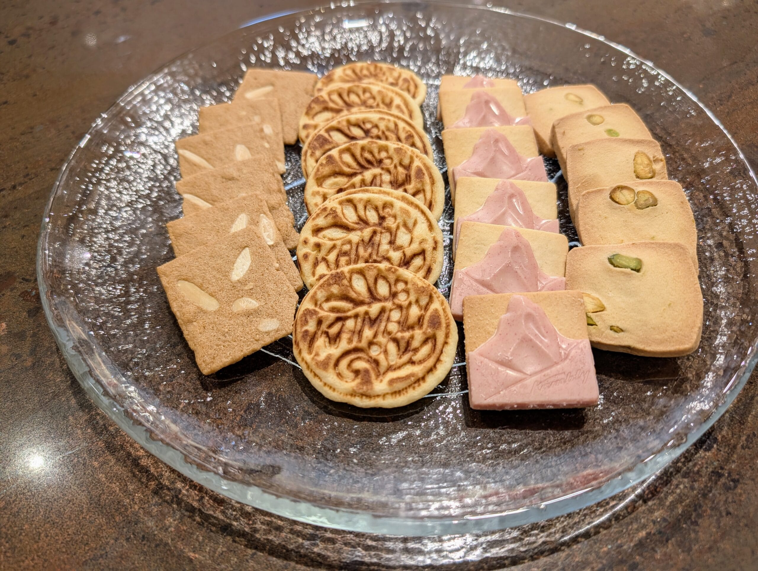 Een glazen schaal met vier verschillende soorten koekjes, waaronder amandelkoekjes, ronde wafeltjes en koekjes met pistache