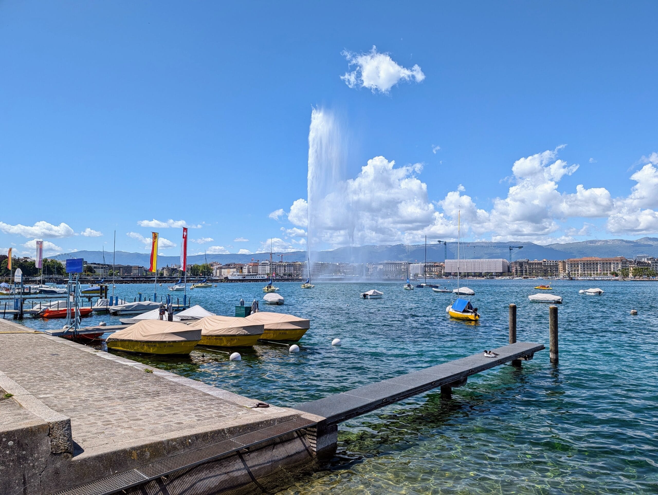 De Jet d'Eau fontein spuit hoog boven het Meer van Genève uit, met boten in de haven en bergen op de achtergrond