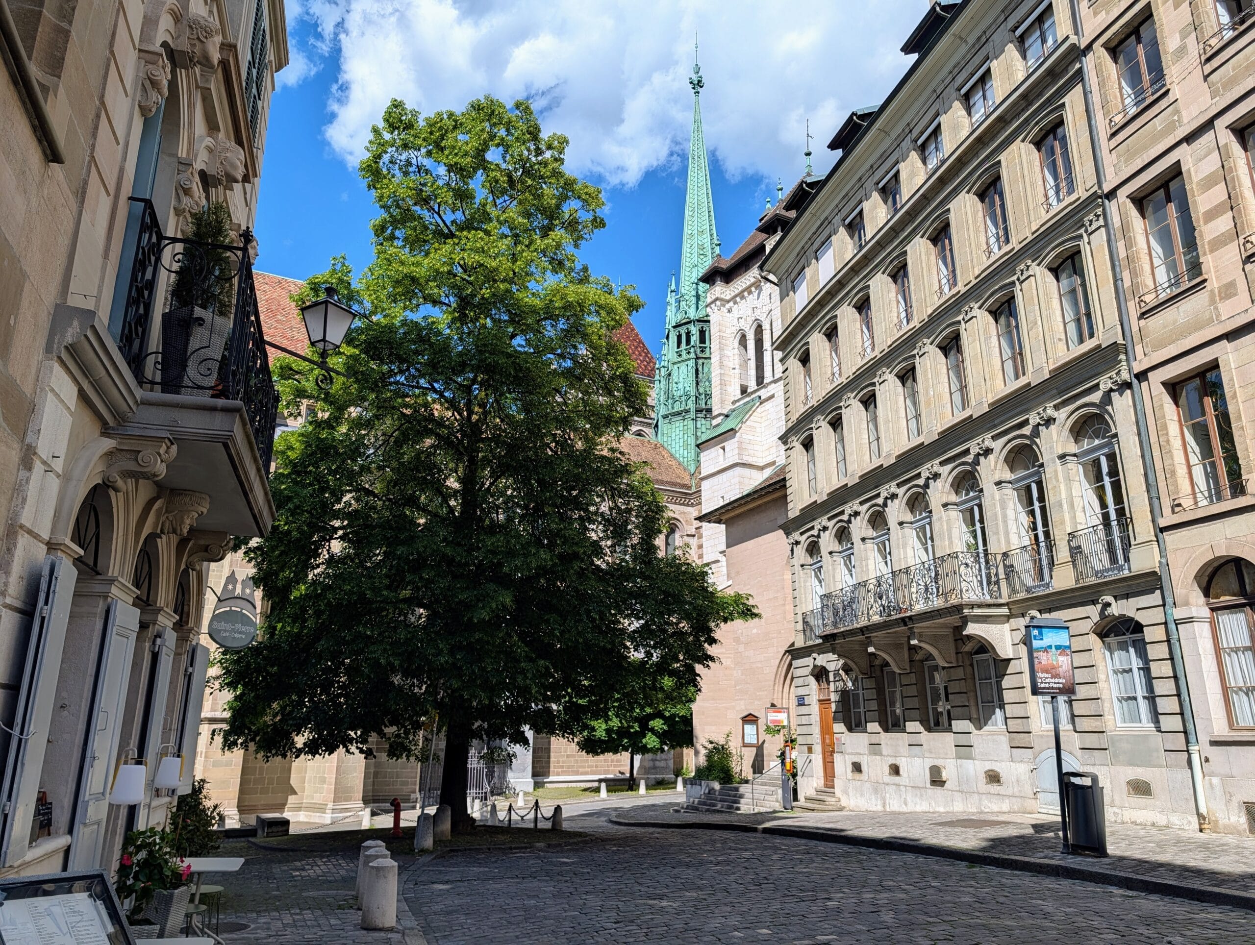 Zicht op een geplaveid plein in Genève met historische gebouwen, een grote boom en de groene spits van de kathedraal