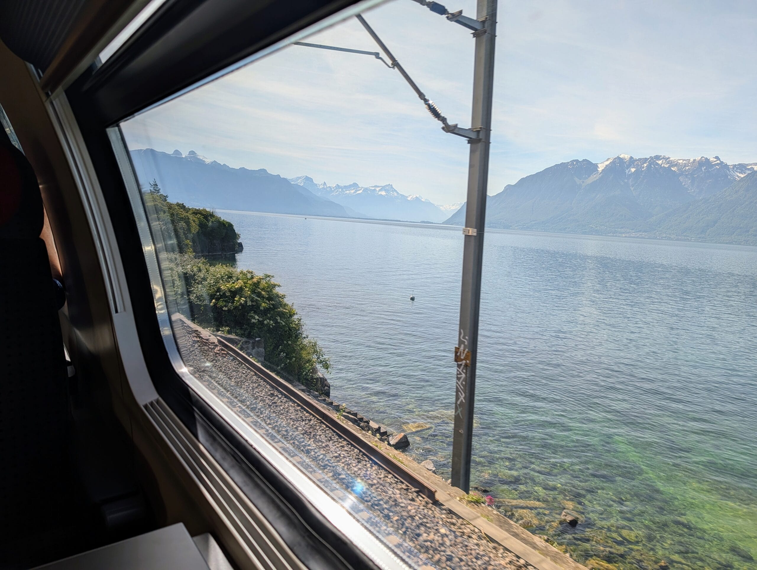 Uitzicht vanuit een treincoupé op een helderblauw meer en besneeuwde bergen onder een lichte hemel