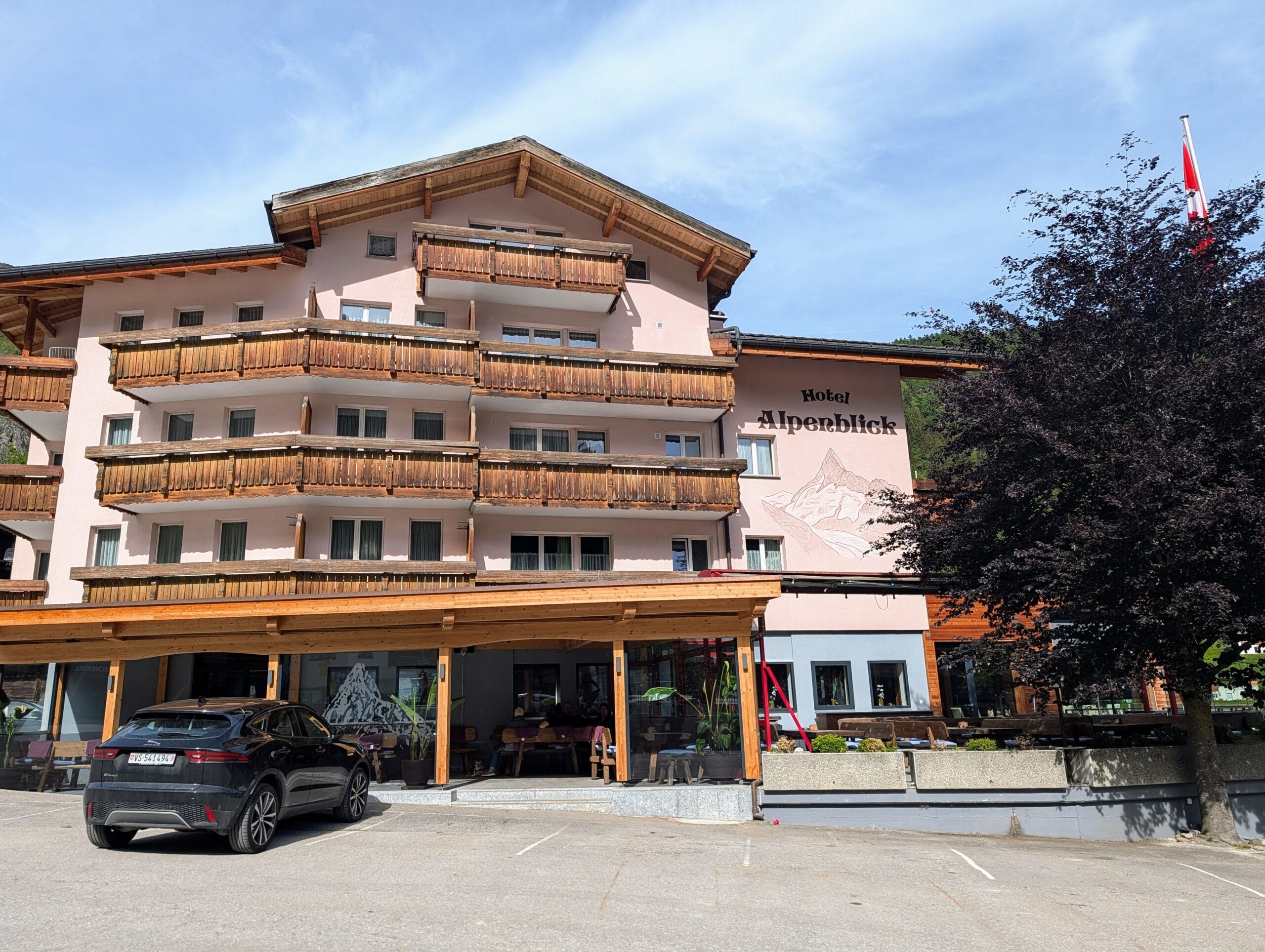 Vooraanzicht van Hotel Alpenblick met houten balkons, een terras en een parkeerplaats met een zwarte auto