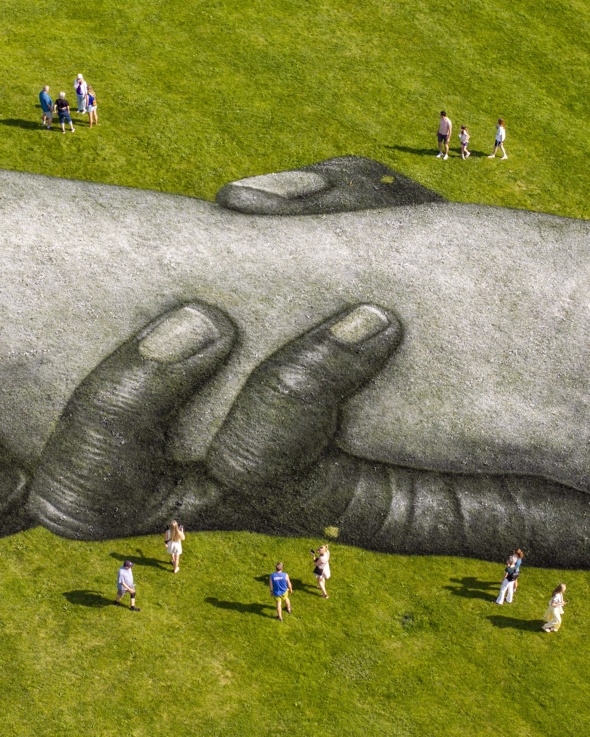 Luchtfoto van een gigantische muurschildering op gras van twee in elkaar grijpende handen, met mensen die eromheen lopen