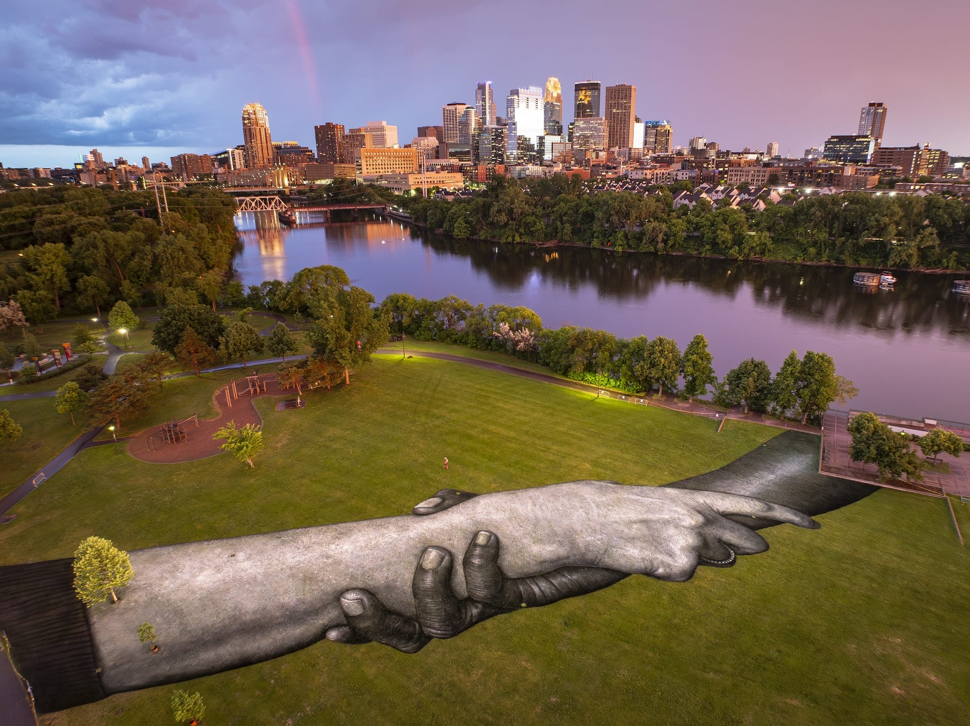 Luchtfoto van een gigantische muurschildering van in elkaar grijpende handen op een grasveld in Minneapolis