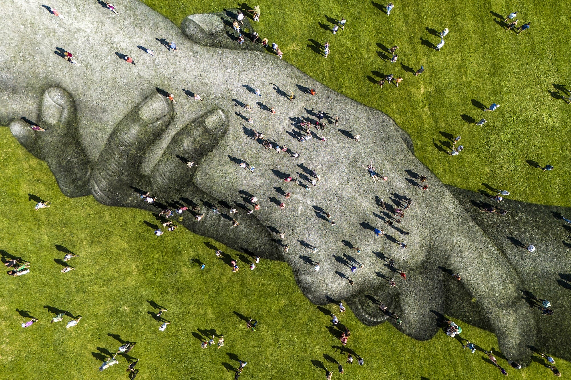 Luchtfoto van een gigantische muurschildering van kunstenaar Saype op gras van twee in elkaar grijpende handen, met mensen die eroverheen lopen