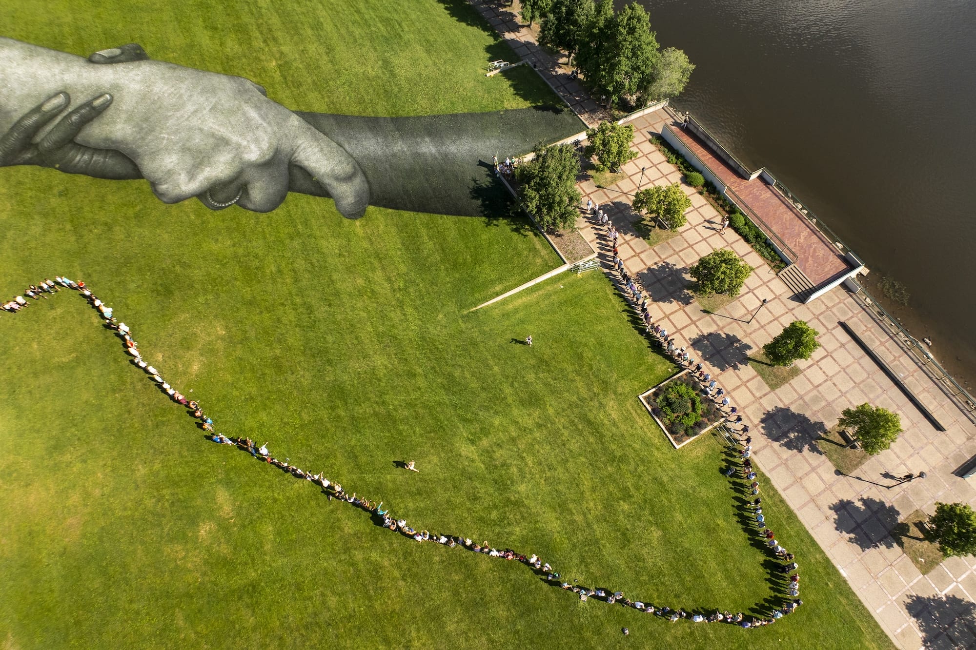 Luchtfoto van een grasveld met een rij mensen en een enorme muurschildering van twee in elkaar grijpende handen