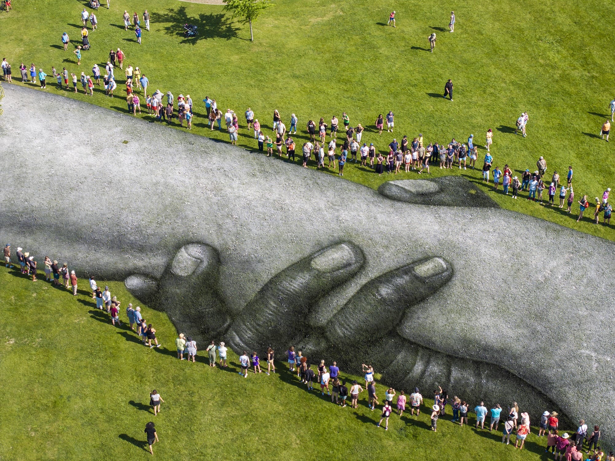 Luchtfoto van een enorm kunstwerk op gras van twee in elkaar grijpende handen, omringd door rijen mensen