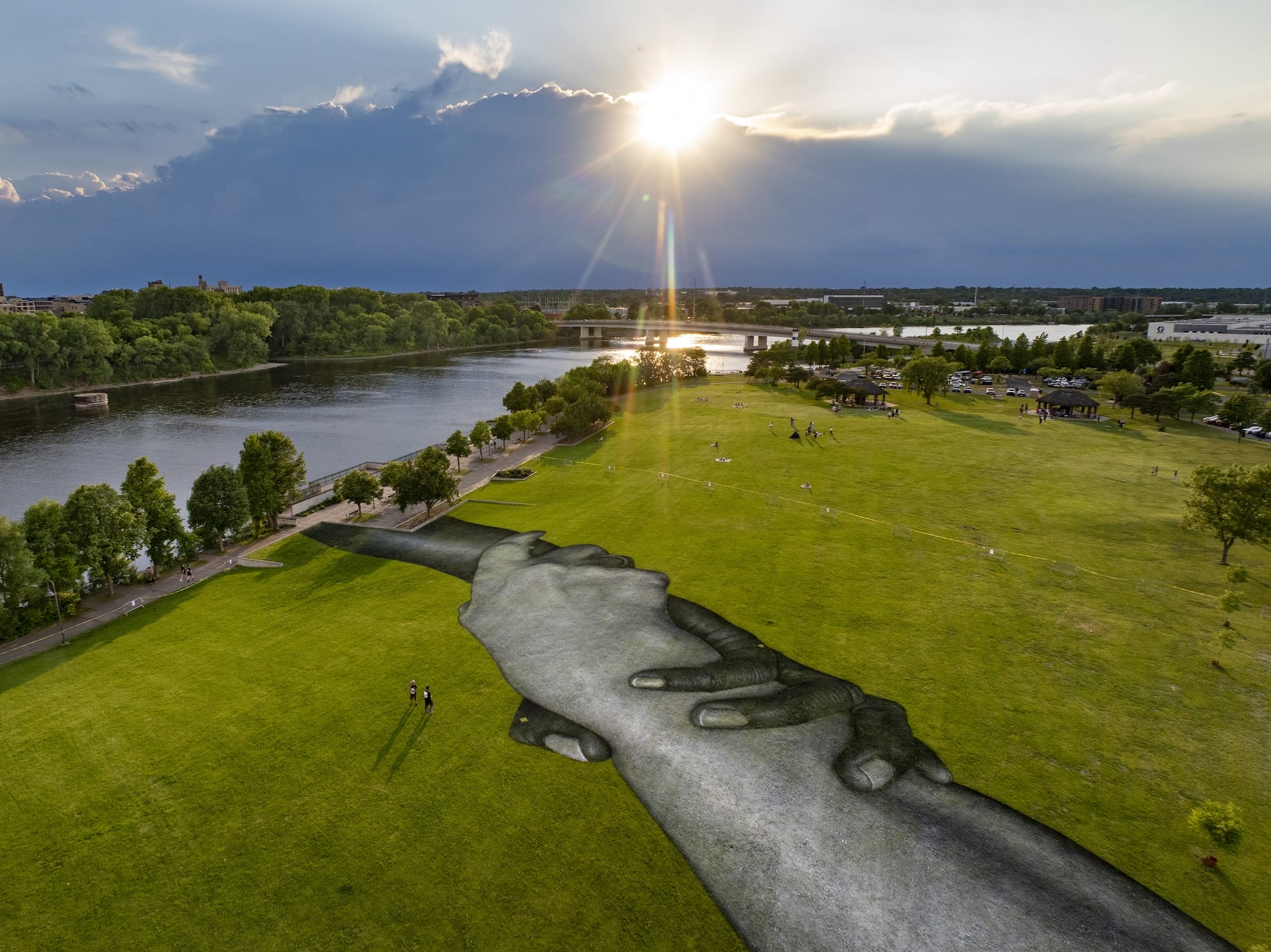 Luchtfoto van een gigantische muurschildering op het gras van een park, die twee in elkaar grijpende handen afbeeldt
