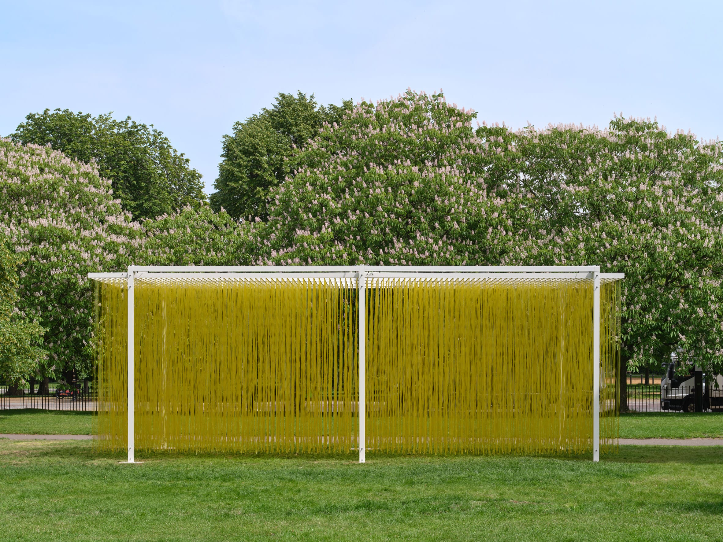 Een wit metalen frame in een park waar honderden gele draden verticaal naar beneden hangen voor een rij bloeiende bomen