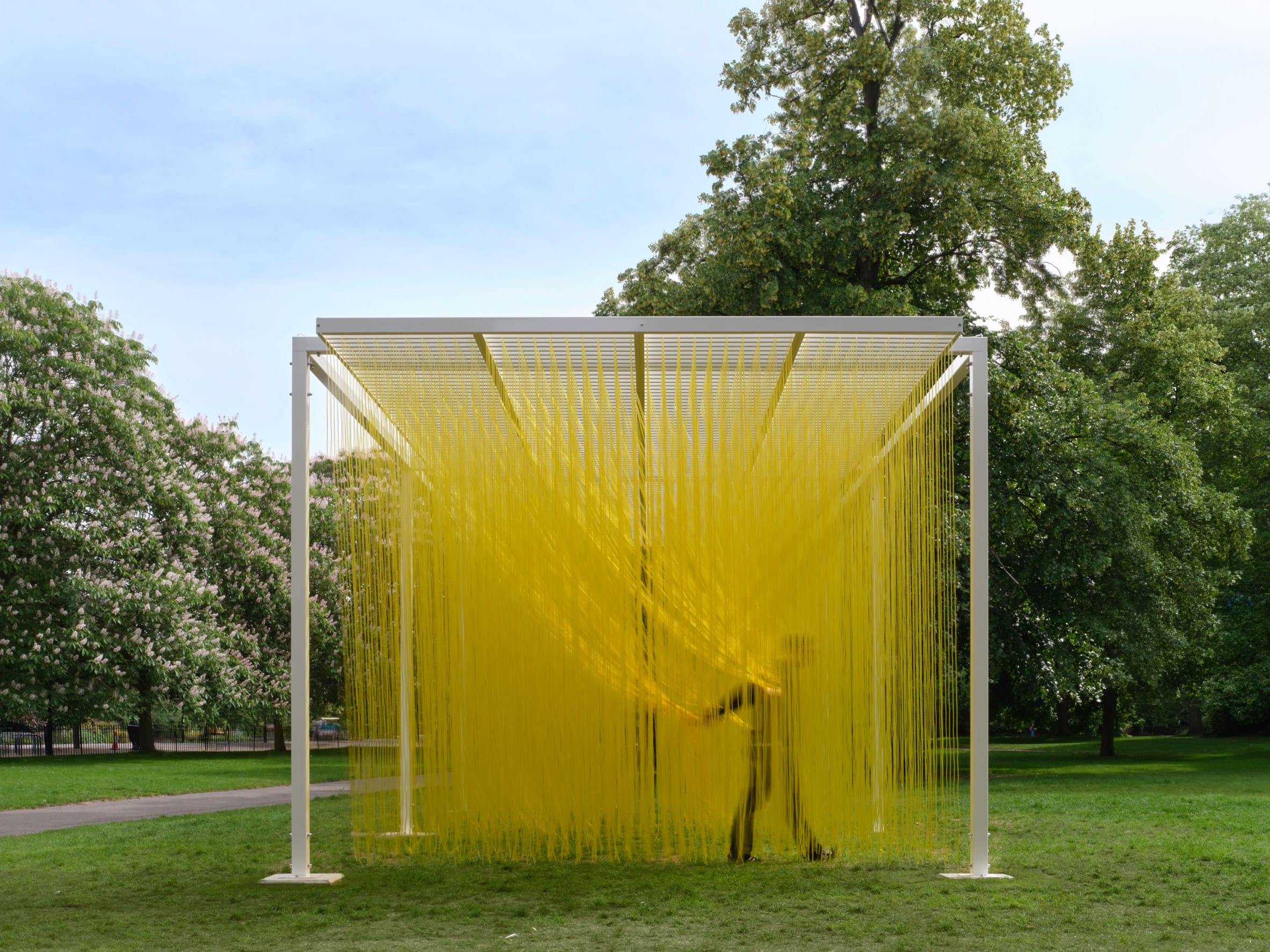Een persoon loopt door een kunstinstallatie van talloze hangende gele draden in een groen park