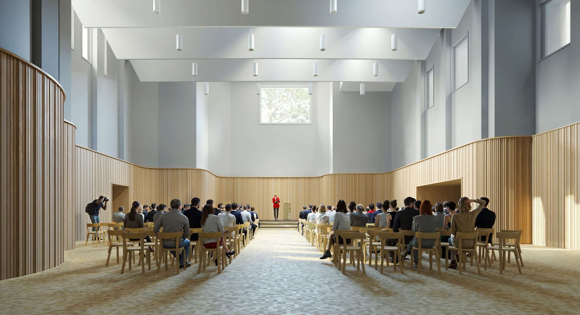 Een spreker in een rood jasje staat voor een publiek in een moderne, lichte zaal met houten wandpanelen