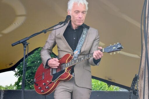 Een man met grijs krullend haar in een beige pak speelt op een rode elektrische gitaar op een podium
