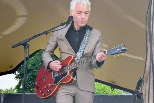 Een man met grijs krullend haar in een beige pak speelt op een rode elektrische gitaar op een podium