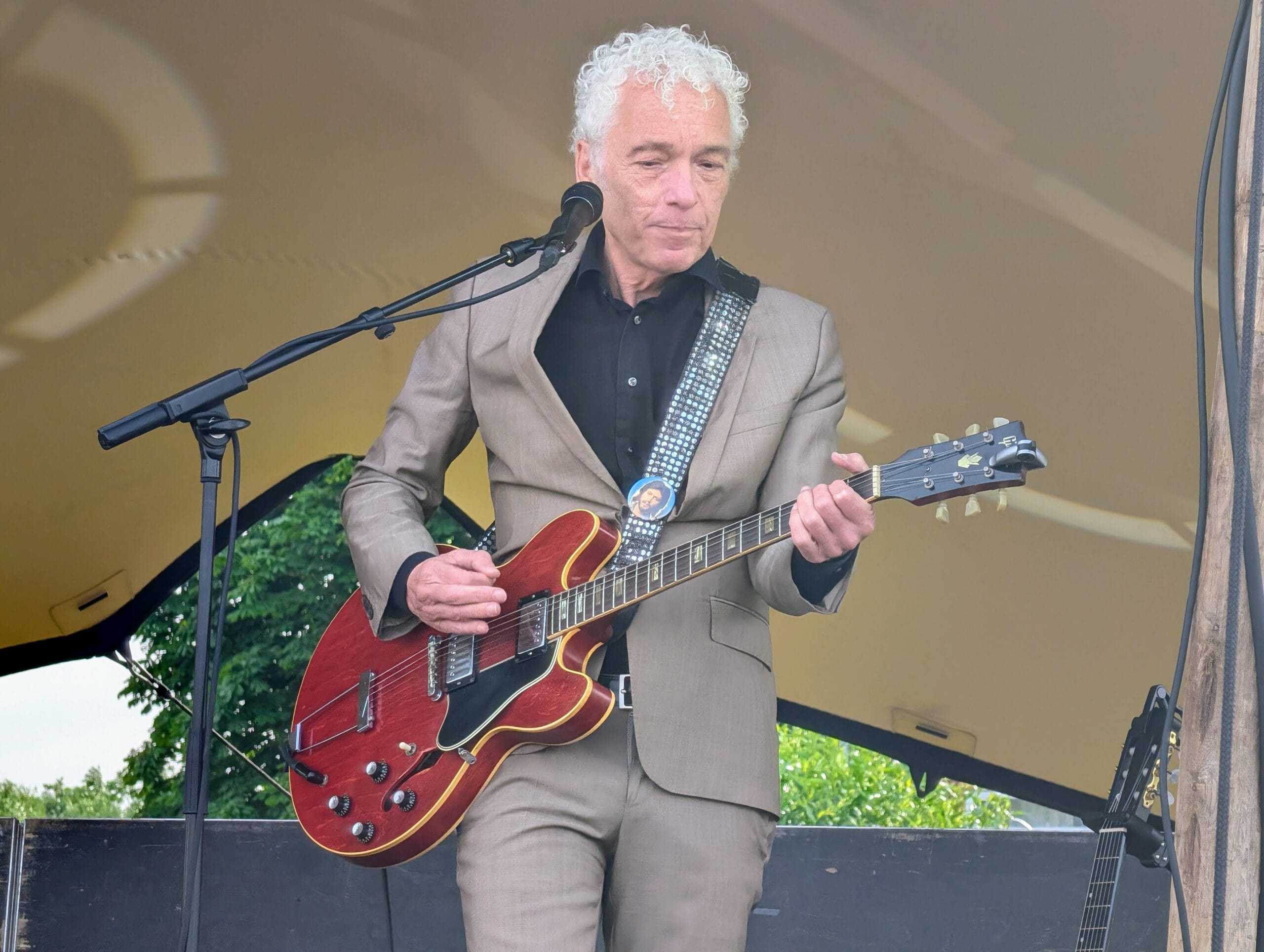 Een man met grijs krullend haar in een beige pak speelt op een rode elektrische gitaar op een podium
