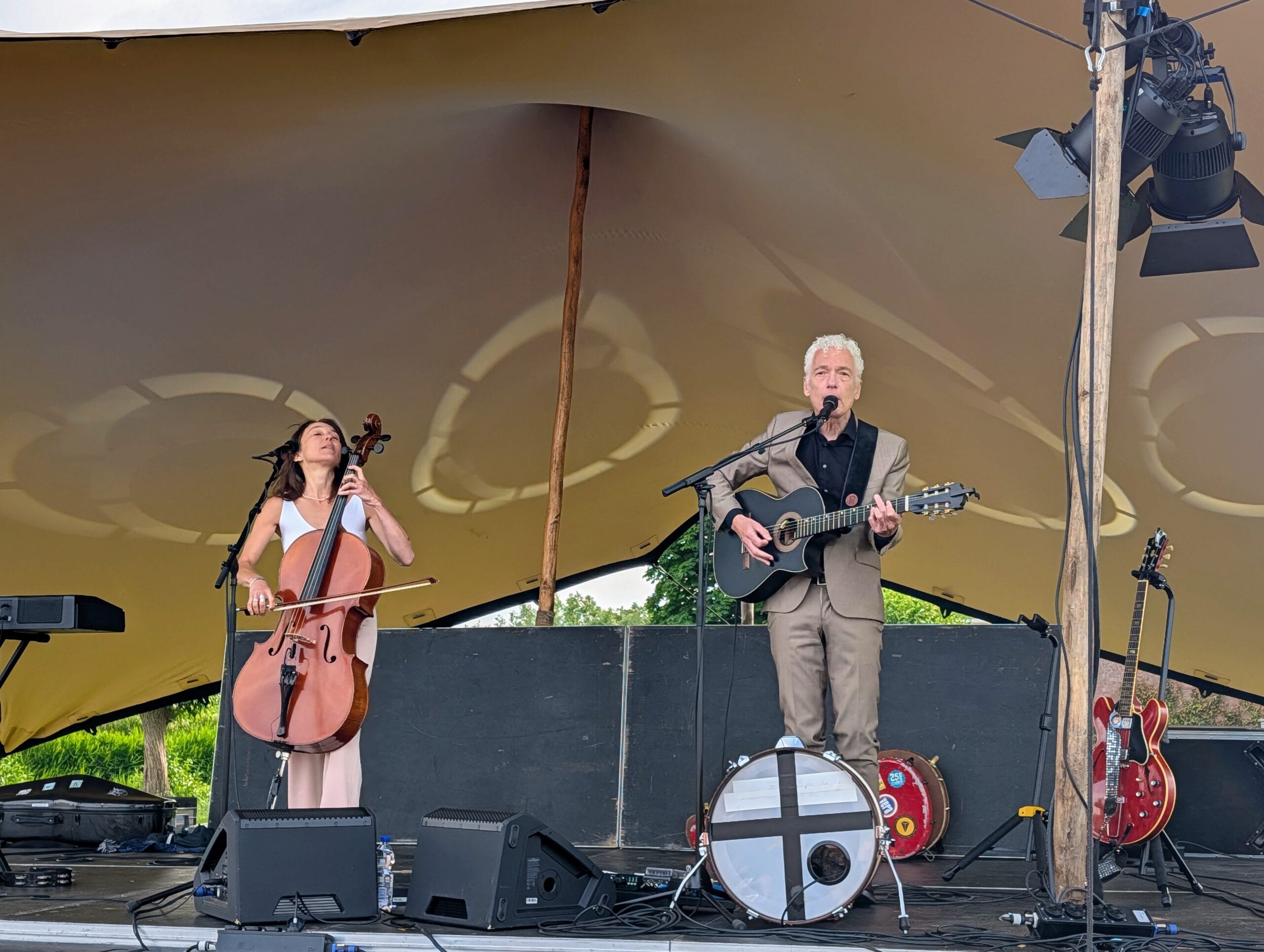 Een man in een beige pak speelt gitaar en zingt op een podium, begeleid door een vrouw op de cello