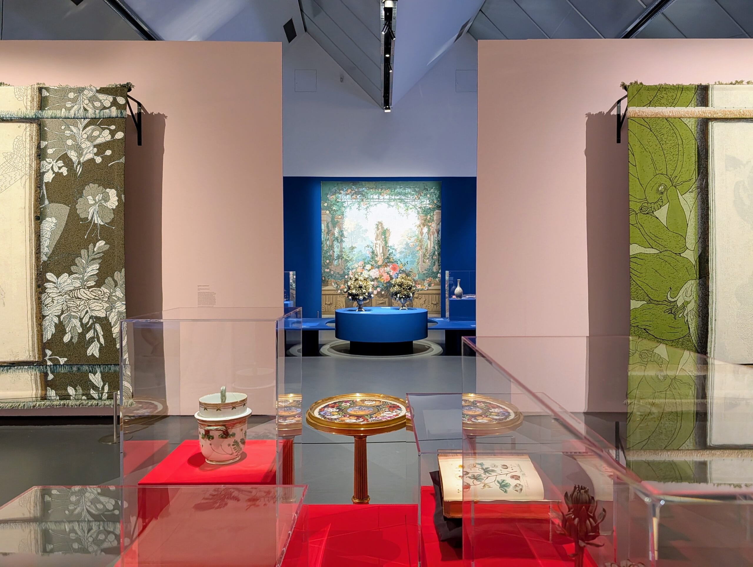 Overzicht van een tentoonstellingsruimte met wandtapijten, porselein en een schilderij van een bloemrijke tuin