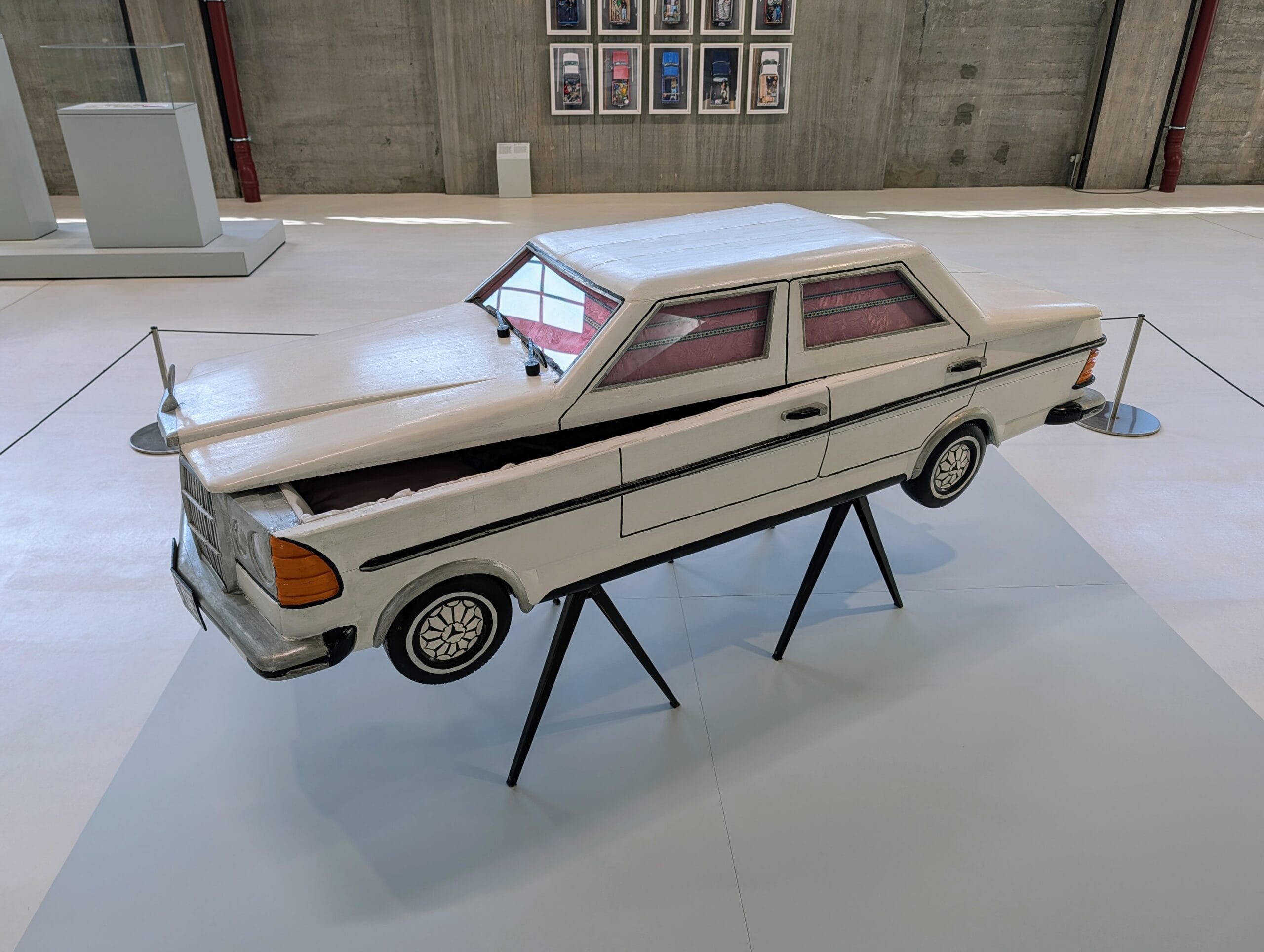 Een wit houten sculptuur van een Mercedes-Benz sedan die als een kist op schragen in een museum staat