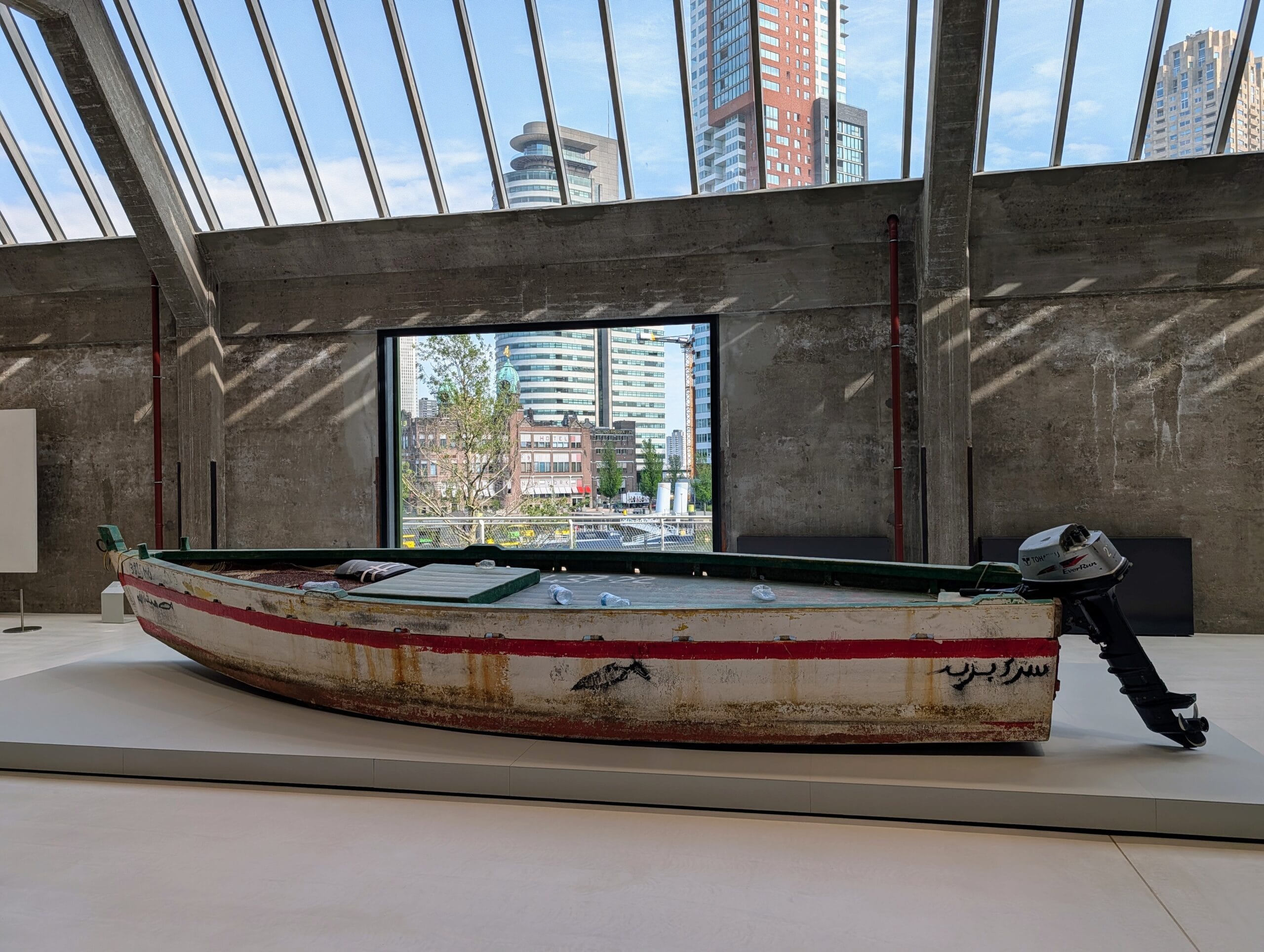 Een verweerde houten boot met een buitenboordmotor tentoongesteld in een museumzaal met uitzicht op de skyline van Rotterdam
