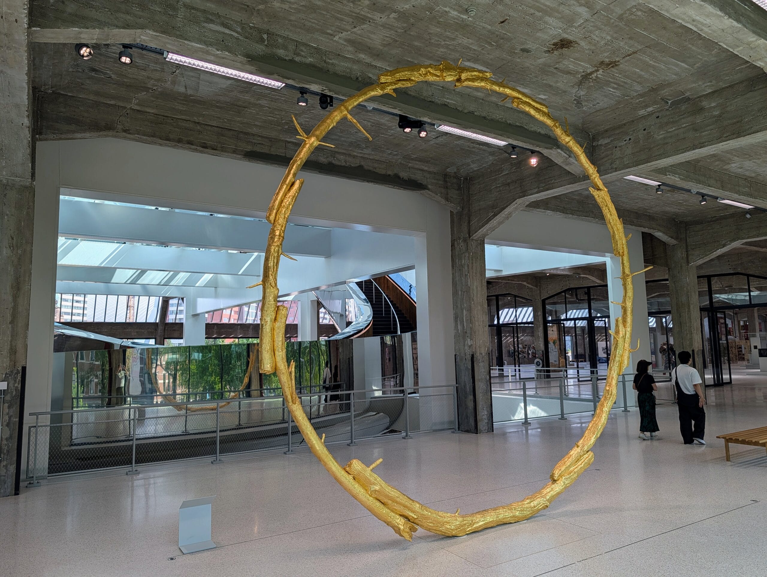 Een grote, goudkleurige cirkelvormige sculptuur van takken in een moderne museumzaal met betonnen kolommen