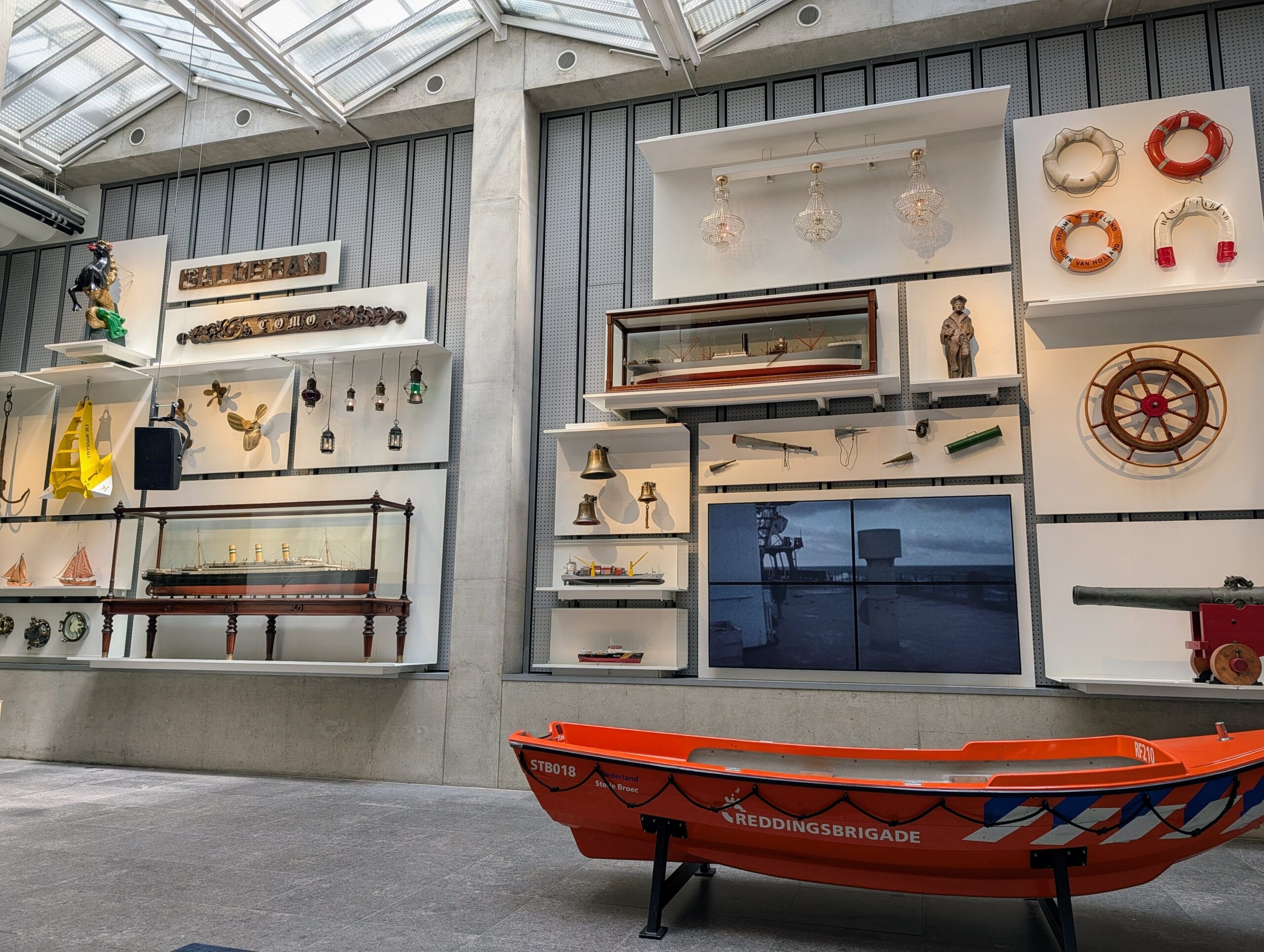 Een museumzaal met maritieme objecten, waaronder scheepsmodellen, reddingsboeien, een stuurwiel en een oranje reddingsboot