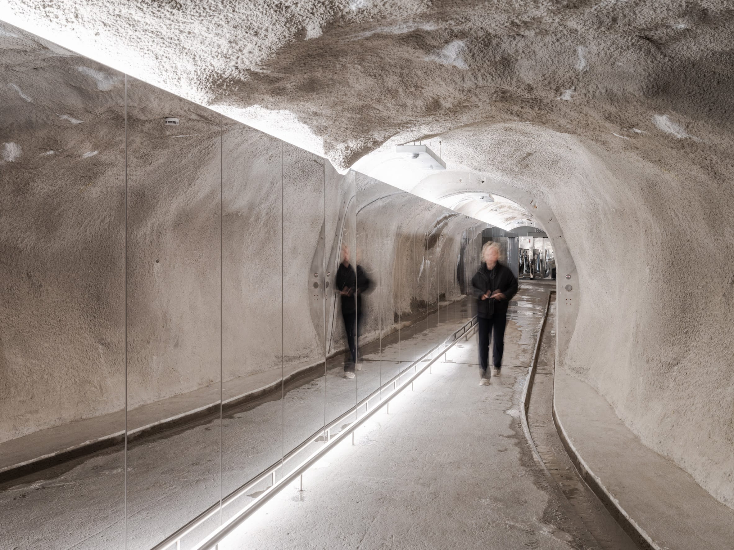 Een persoon loopt door een verlichte, moderne tunnel met ruwe betonnen wanden en een grote spiegelwand