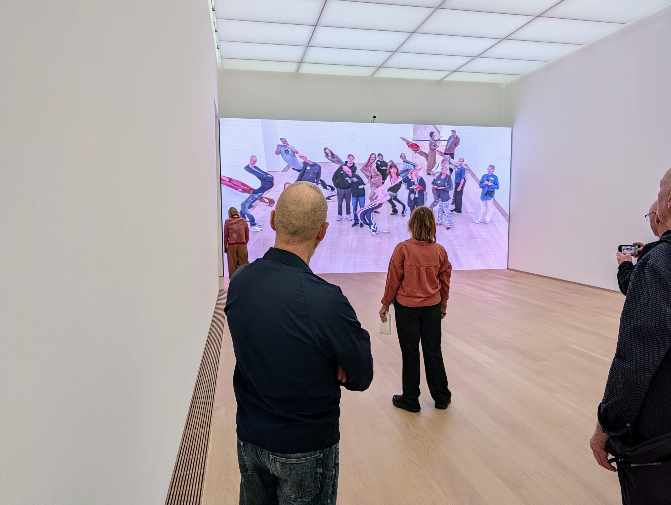 Bezoekers in een museum bekijken een groot scherm waarop een vervormde groepsfoto van mensen wordt getoond