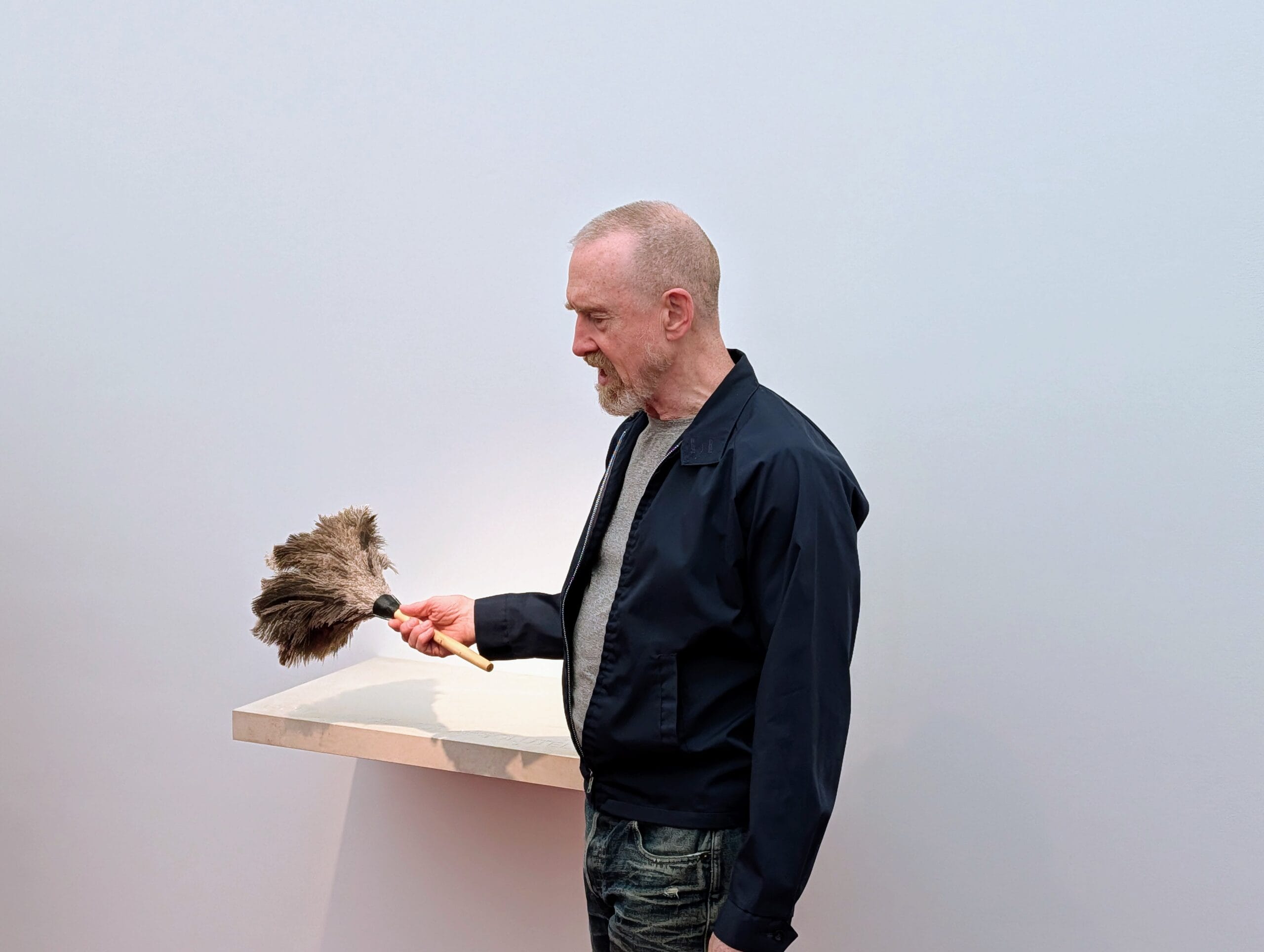 Een man met een baard houdt een plumeau vast boven een witte plank tegen een lichte achtergrond