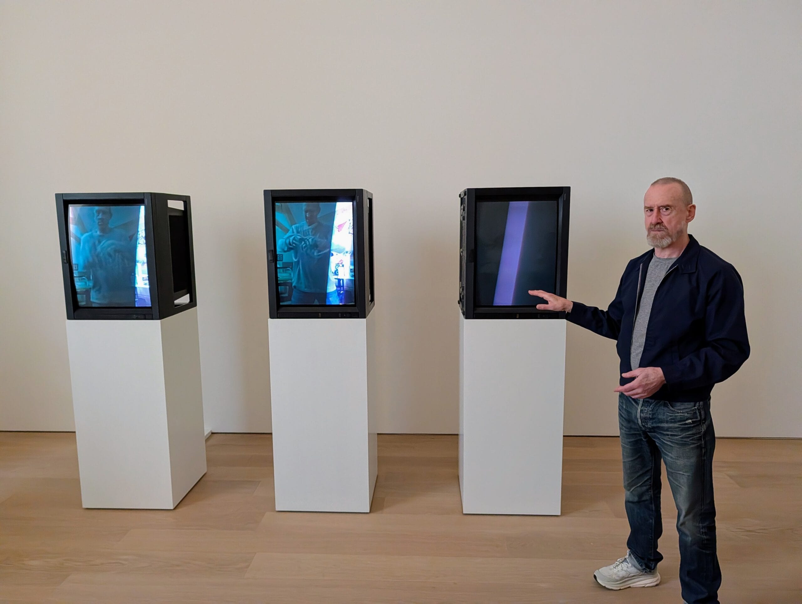 Een man staat naast drie monitoren op witte zuilen in een galerij en wijst naar het rechter scherm