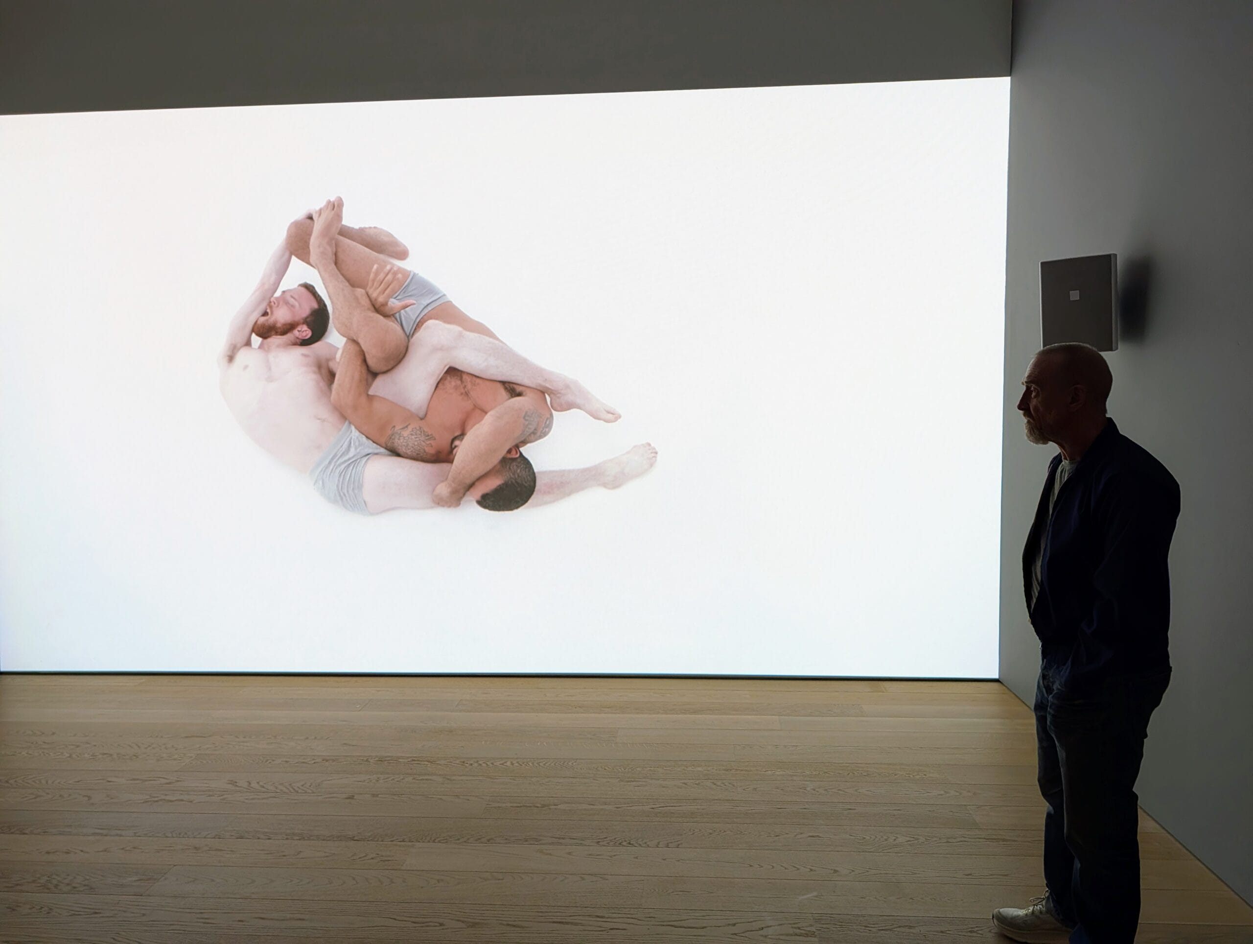 Een man in een museum bekijkt een grote videoprojectie van twee worstelende mannen in ondergoed tegen een witte achtergrond