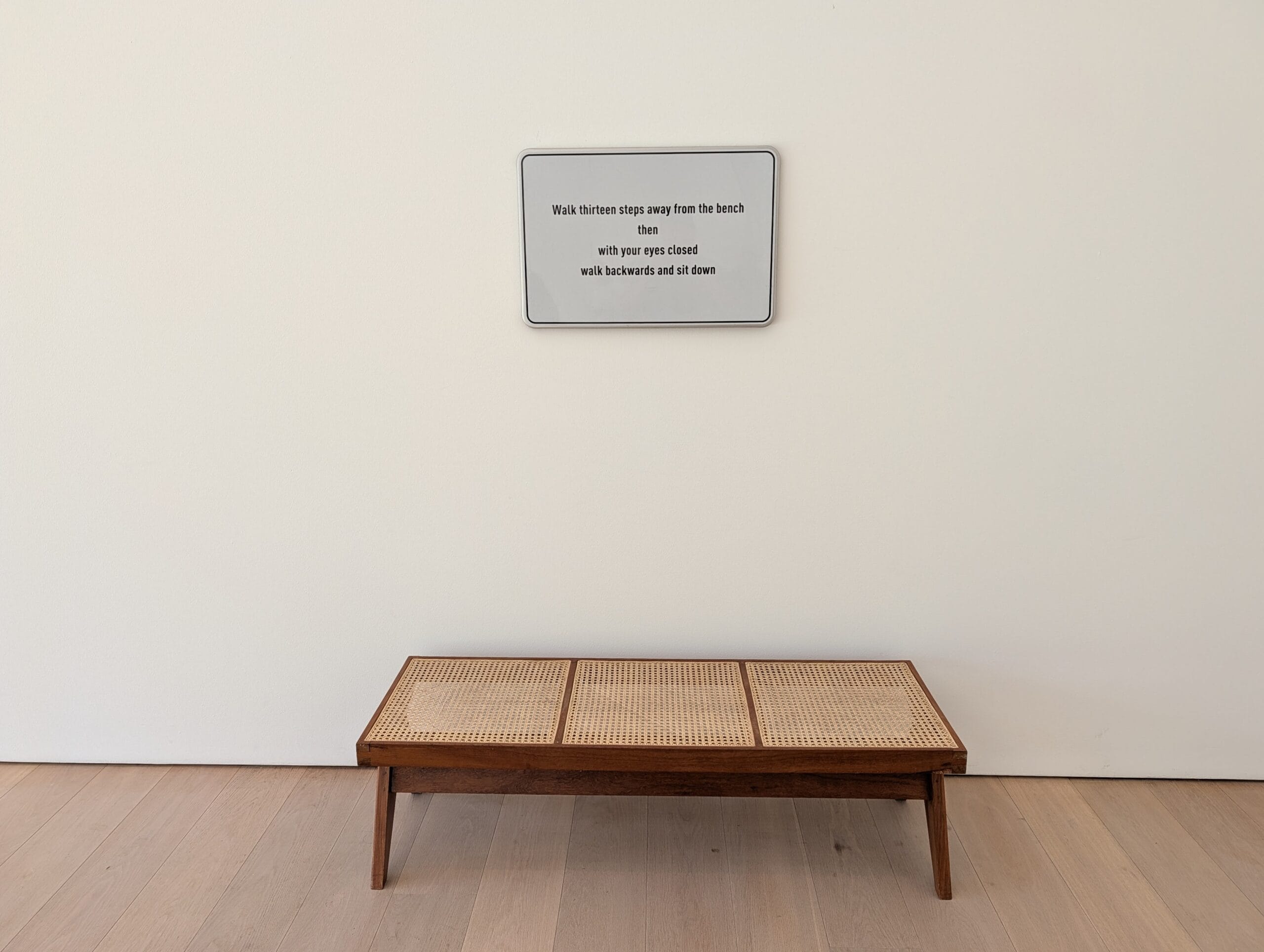 Een houten bankje met een rieten zitting voor een witte muur met een bordje met instructies voor een kunstwerk