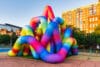 Een groot, opblaasbaar kunstwerk bestaande uit in elkaar verstrengelde, kleurrijke buizen in regenboogkleuren op een plein