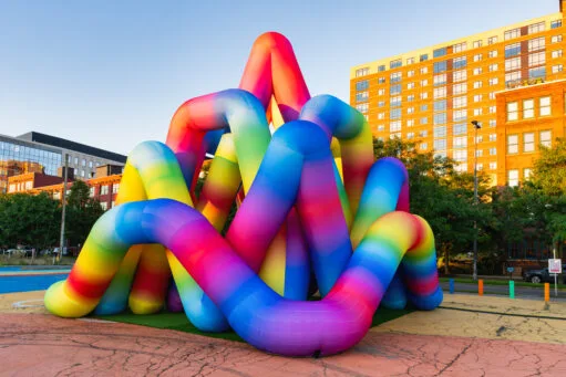Een groot, opblaasbaar kunstwerk bestaande uit in elkaar verstrengelde, kleurrijke buizen in regenboogkleuren op een plein