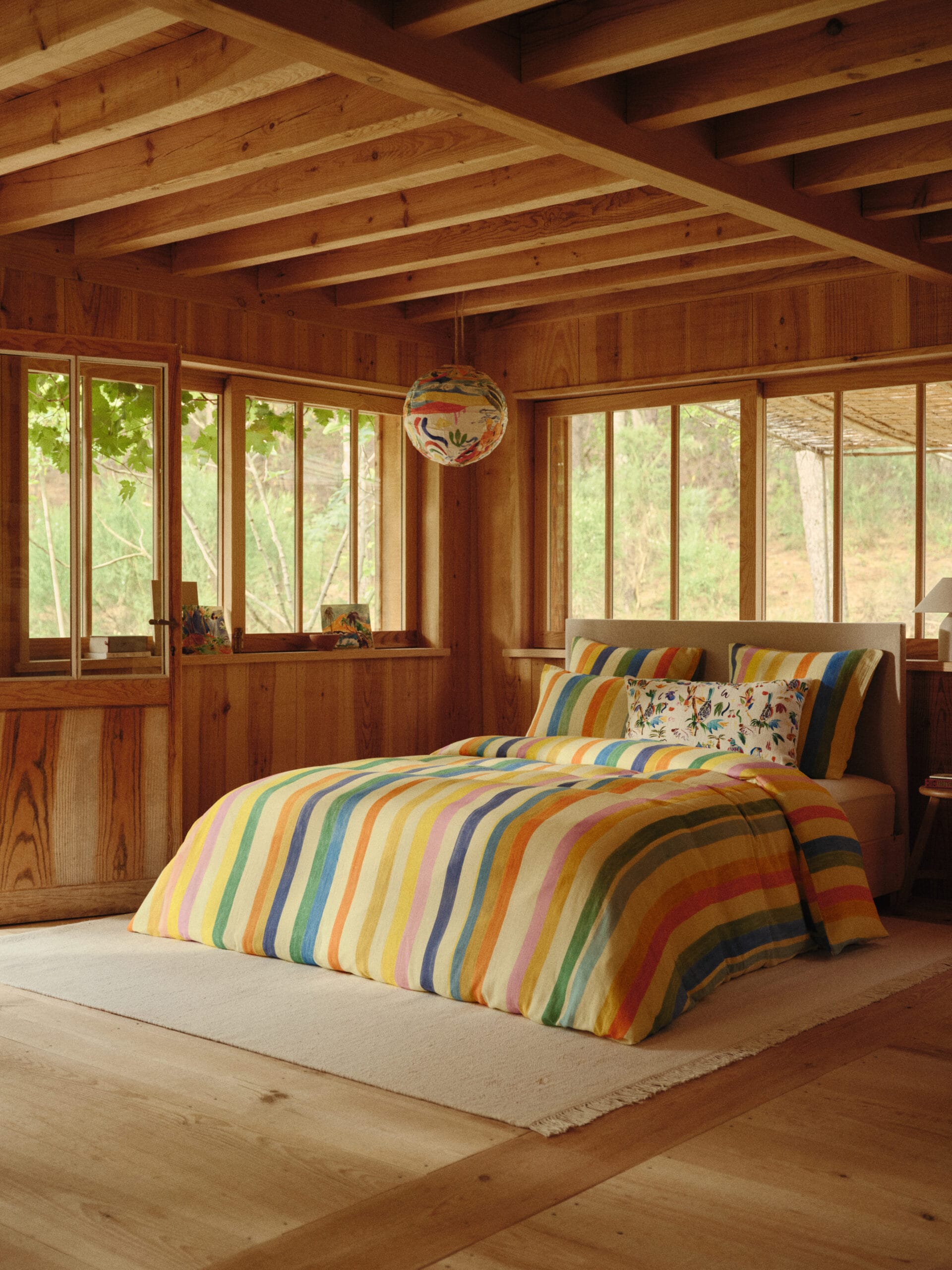 Slaapkamer met een houten wanden en plafond met een bed opgemaakt in kleurrijk gestreept beddengoed en een bijpassende lampion, uit de gezamenlijke collectie van Bonsoirs en G.Kero