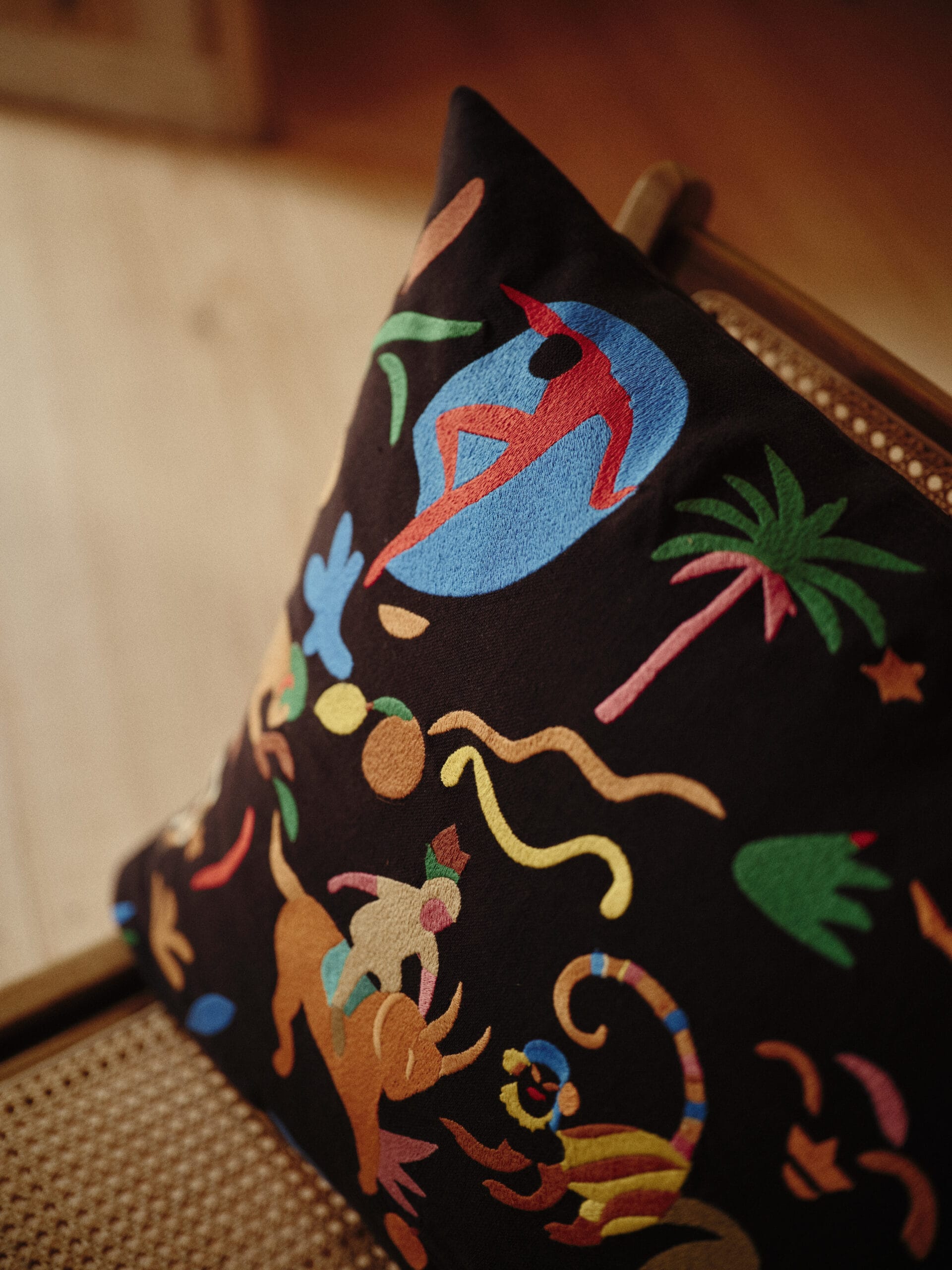 Zwart kussen met kleurrijk borduursel van een tropisch tafereel met figuren, dieren en een palmboom, uit de gezamenlijke collectie van Bonsoirs en G.Kero