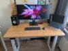 Een houten zit-sta bureau met een breed beeldscherm, luidsprekers, een laptop op een standaard en een gitaar op de achtergrond