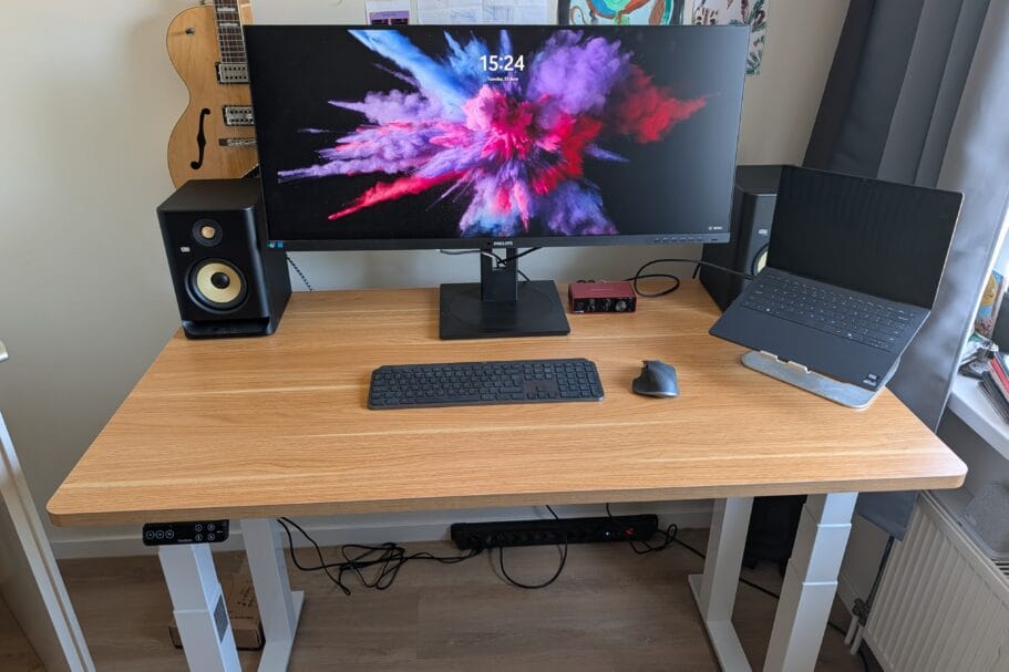 Een houten zit-sta bureau met een breed beeldscherm, luidsprekers, een laptop op een standaard en een gitaar op de achtergrond
