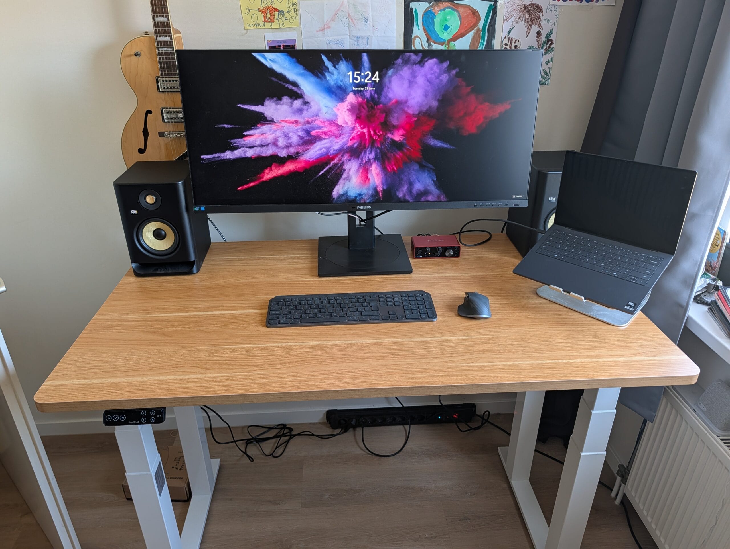 Een houten zit-sta bureau met een breed beeldscherm, luidsprekers, een laptop op een standaard en een gitaar op de achtergrond
