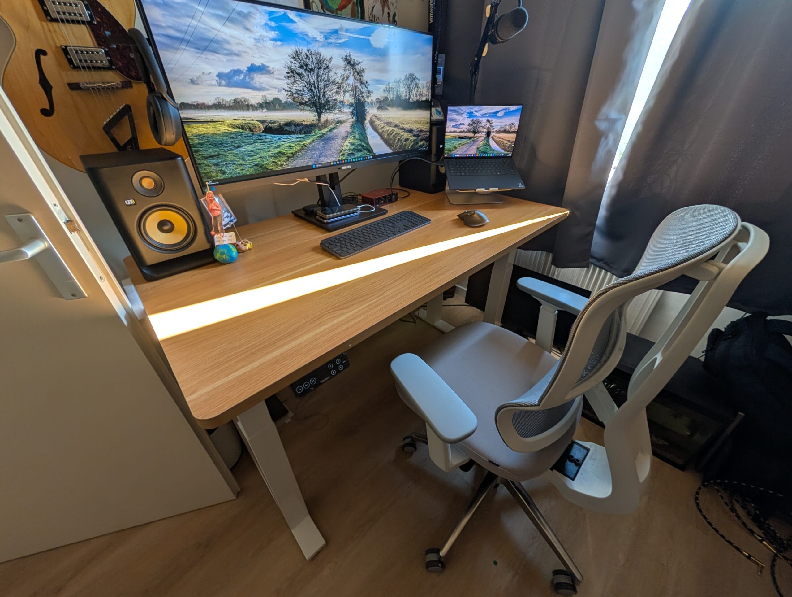 Een modern thuiskantoor met een houten zit-sta bureau, een grote monitor, laptop, ergonomische bureaustoel en een gitaar