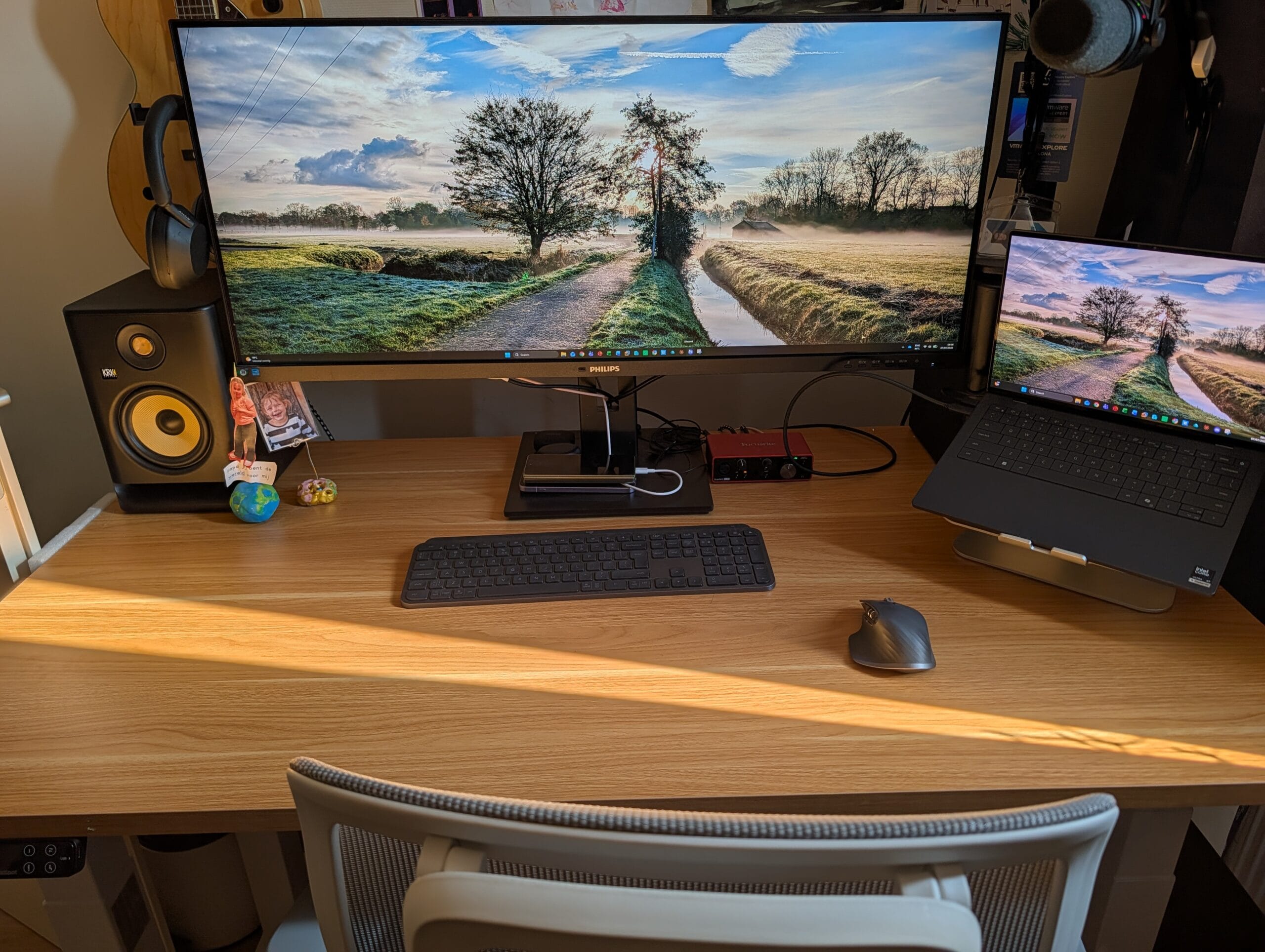 Een houten bureau met een breed Philips-scherm, een laptop op een standaard, een toetsenbord, muis en studiomonitoren