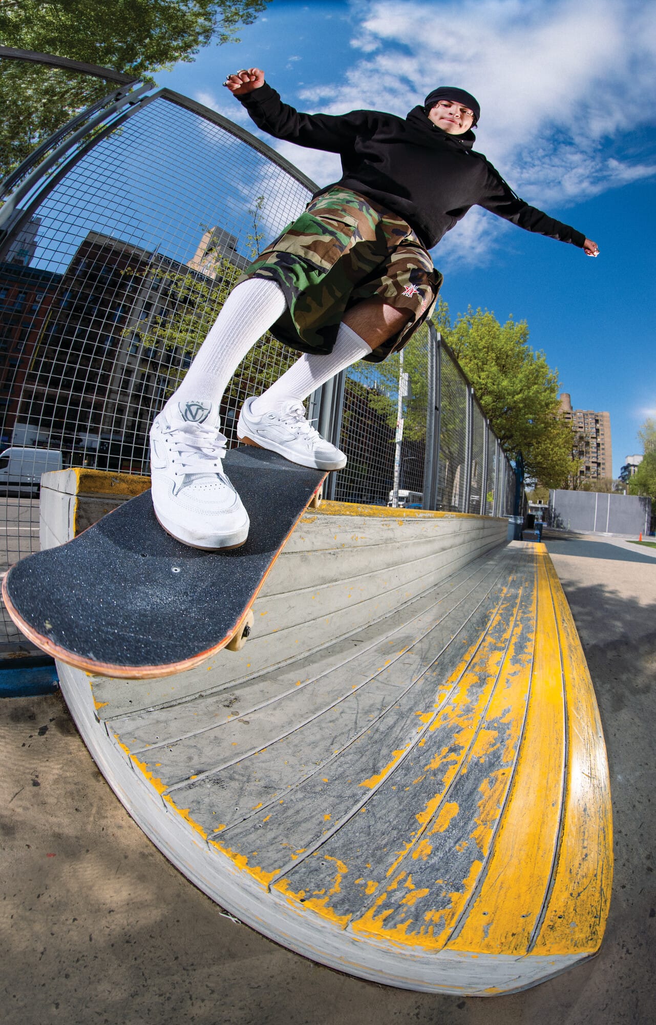 Een skateboarder in een zwarte hoodie en camouflagebroek voert een trick uit op een betonnen rand in een skatepark