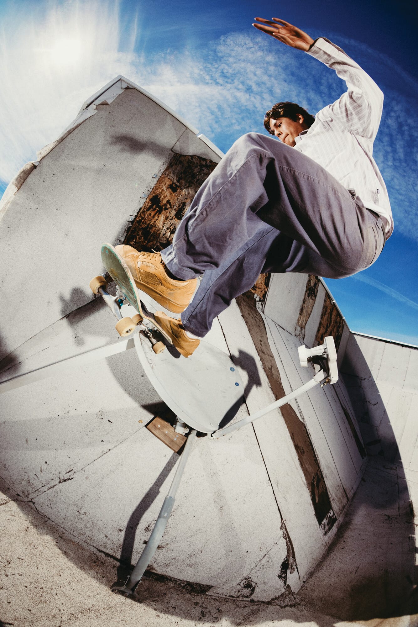 Een skateboarder voert een sprong uit over een schotelantenne op een betonnen dak tegen een blauwe lucht