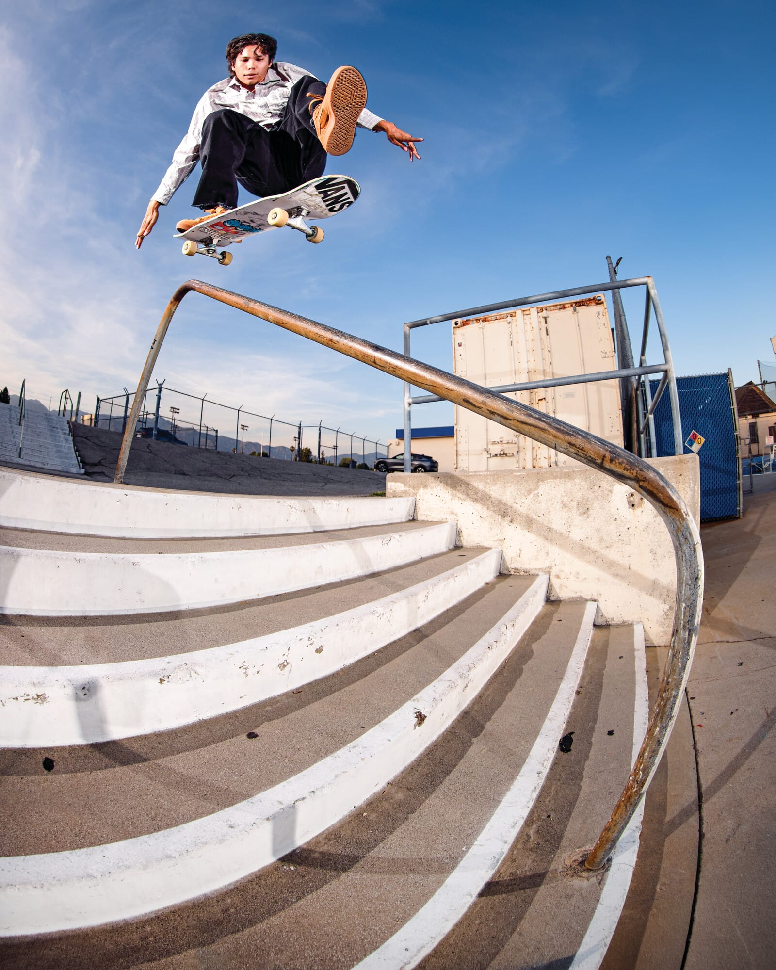 Een skateboarder voert een hoge sprong uit over een trapleuning tegen een blauwe lucht