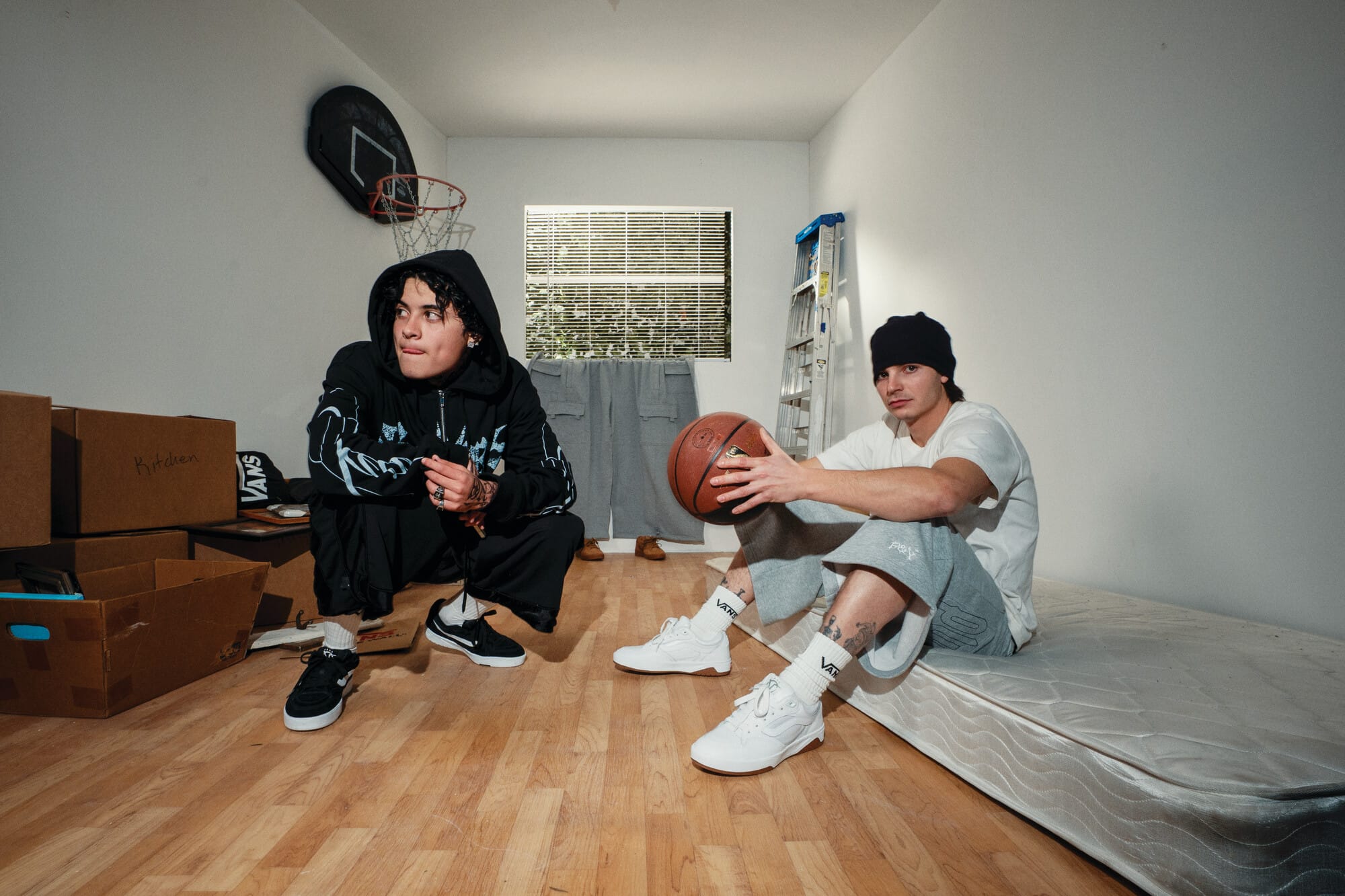 Twee jonge mannen in streetwear poseren in een kamer met verhuisdozen, een basketbal en een matras op de vloer