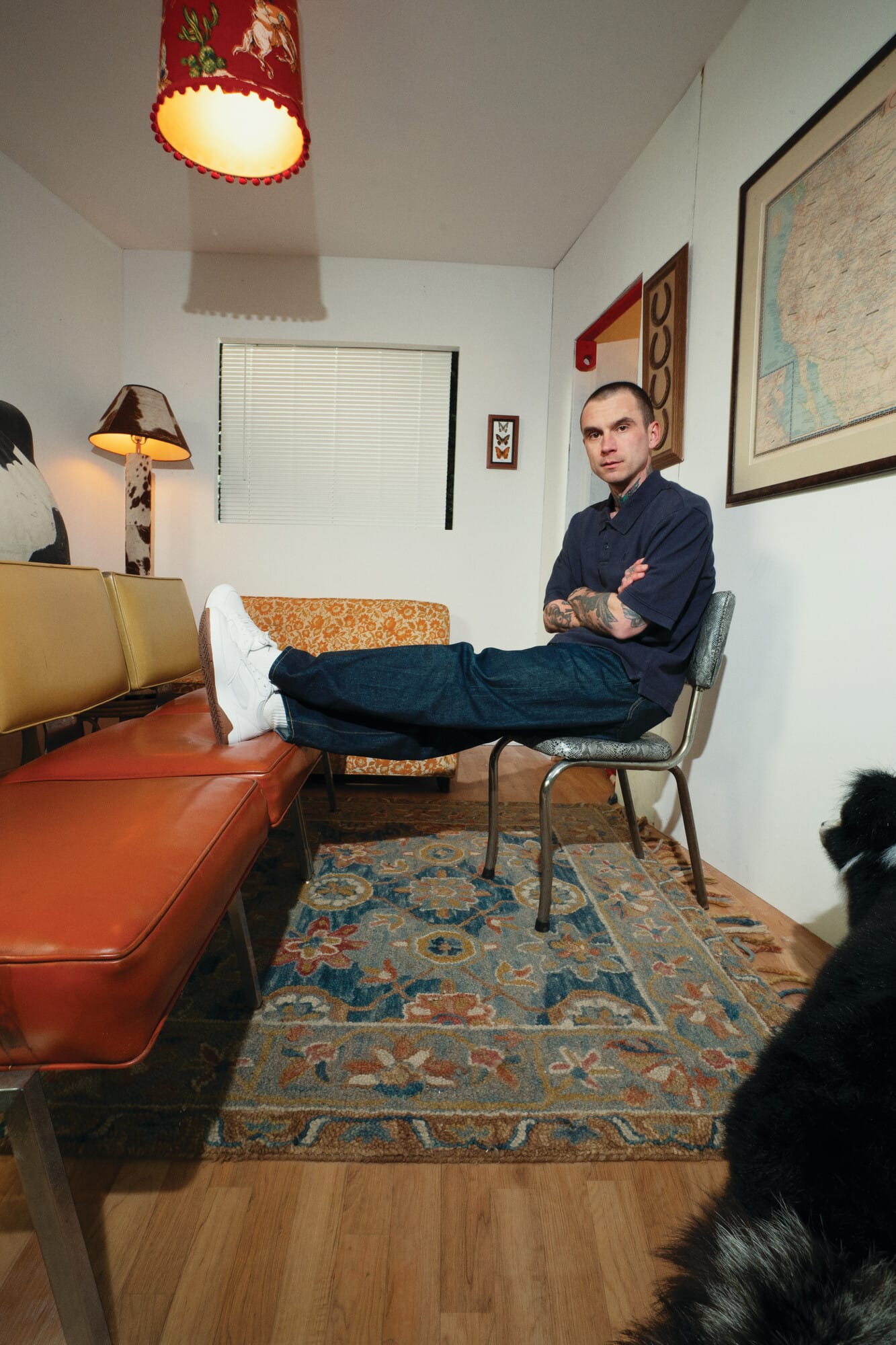 Een man met getatoeëerde armen zit op een stoel met zijn voeten op een oranje bank in een eclectisch ingerichte kamer