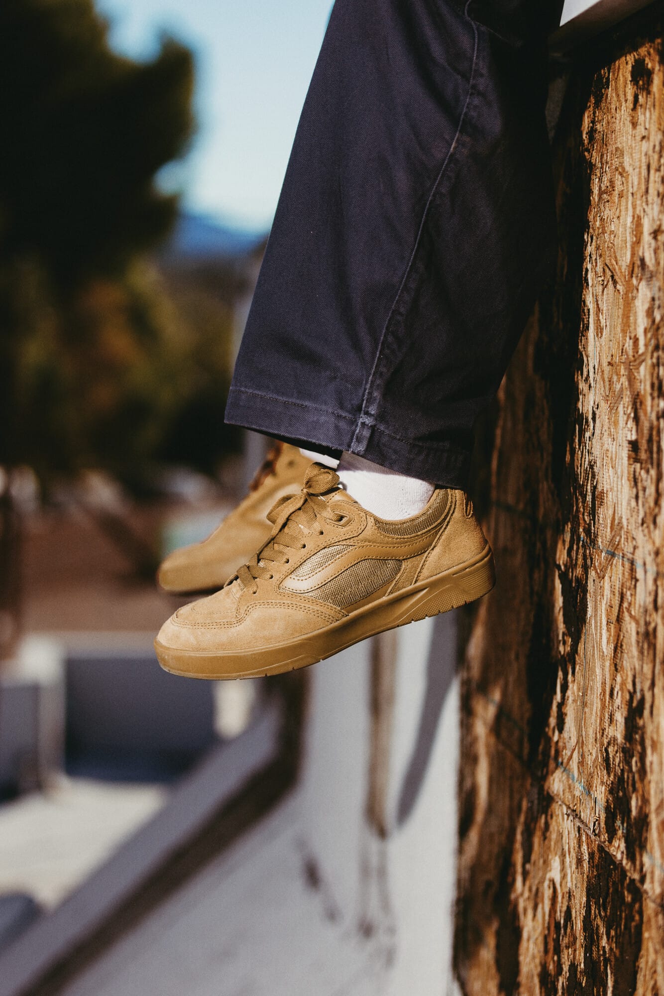 Iemand die op een houten rand zit met bruine suède sneakers en een donkerblauwe broek